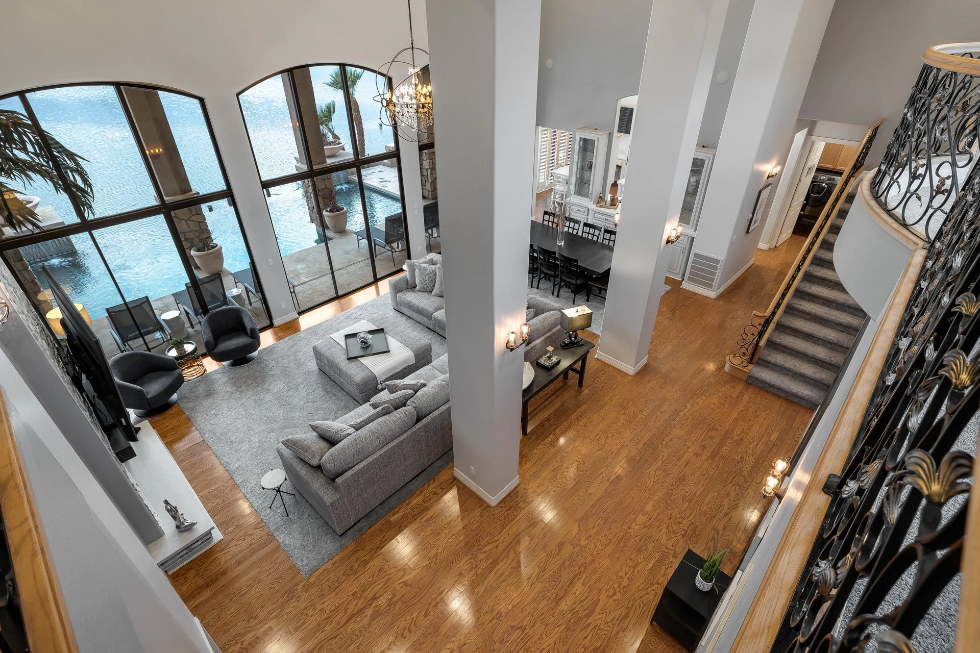 Living room with large windows overlooking water, furnished with gray sofas, black armchairs, a gray rug, and a glass-top coffee table. Adjacent dining area has a long black table with eight chairs, white cabinets, and a chandelier. Hardwood floors, 