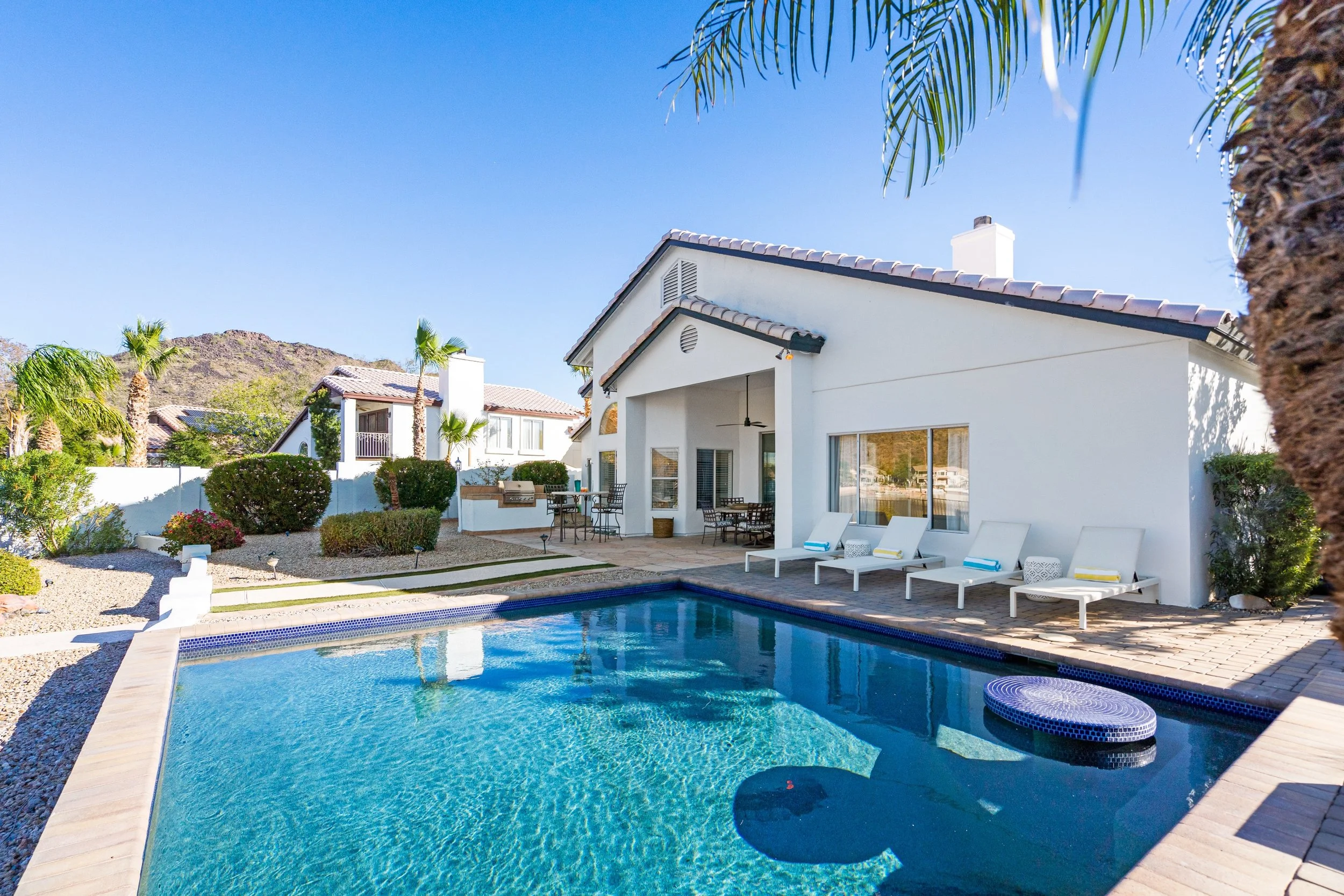 Backyard view of a white house with a swimming pool, lounge chairs, and outdoor seating area under a clear blue sky, with palm trees and neighboring houses in the background.