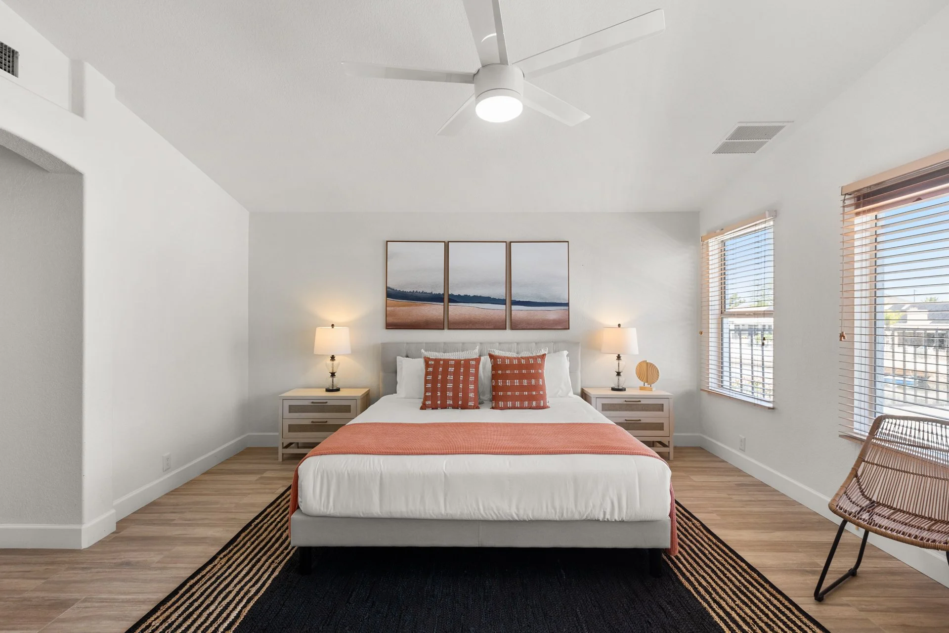 A modern bedroom with a white bed, orange accents, two nightstands with lamps, a framed landscape artwork, and large windows with blinds letting in natural light.