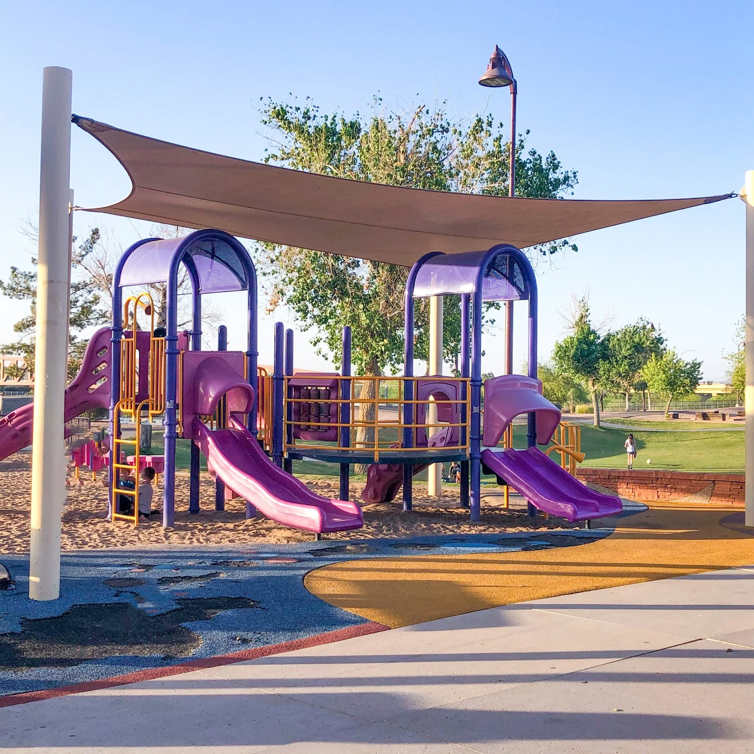 A colorful playground at a park with slides, climbing structures, and shade canopies, surrounded by trees and a clear blue sky.