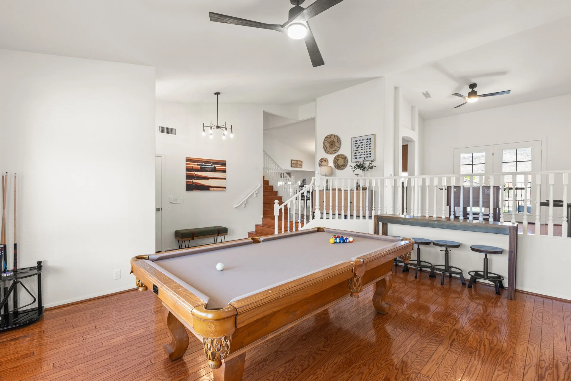 Living room with a pool table, hardwood floors, ceiling fans, and a staircase leading to an upper level with large windows, bar stools, and wall decor.