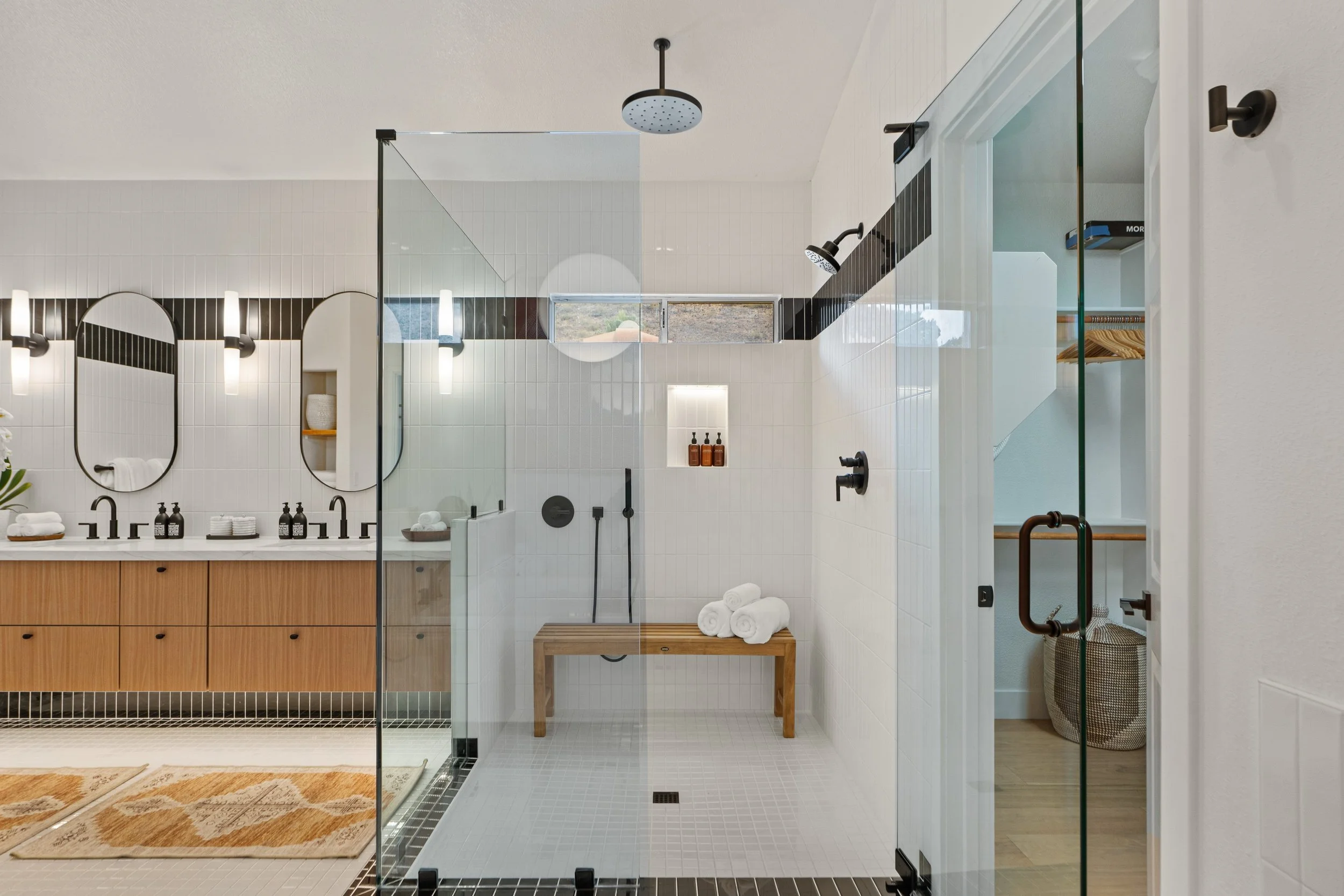 Modern bathroom with a glass shower enclosure, white tiled walls with black accents, dual mirrors above a wooden vanity, and black fixtures for the sinks and shower.