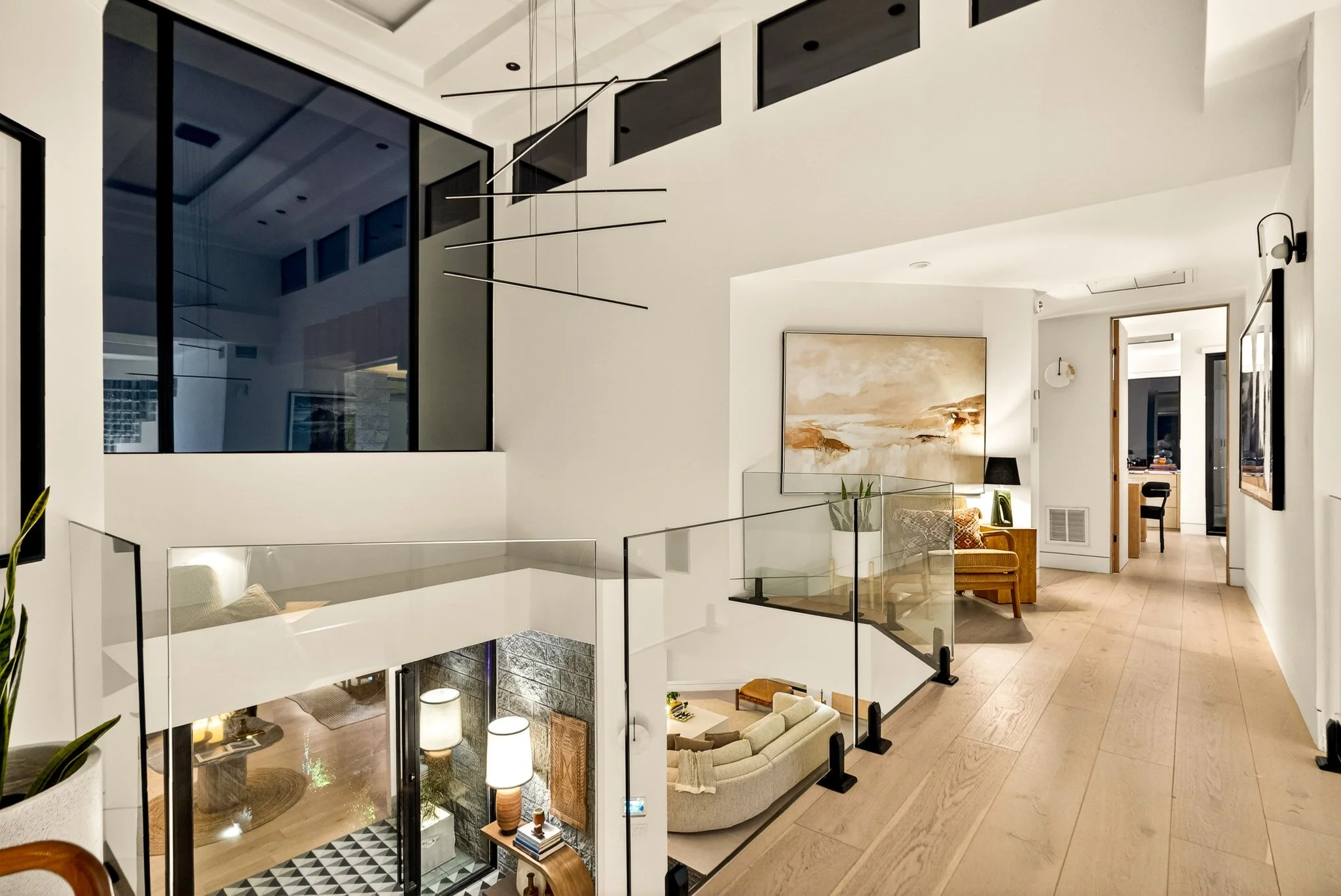 Interior view of a modern multi-level home with open floor plan, featuring white walls, large windows, a black metal staircase with glass railing, and artwork on the walls.