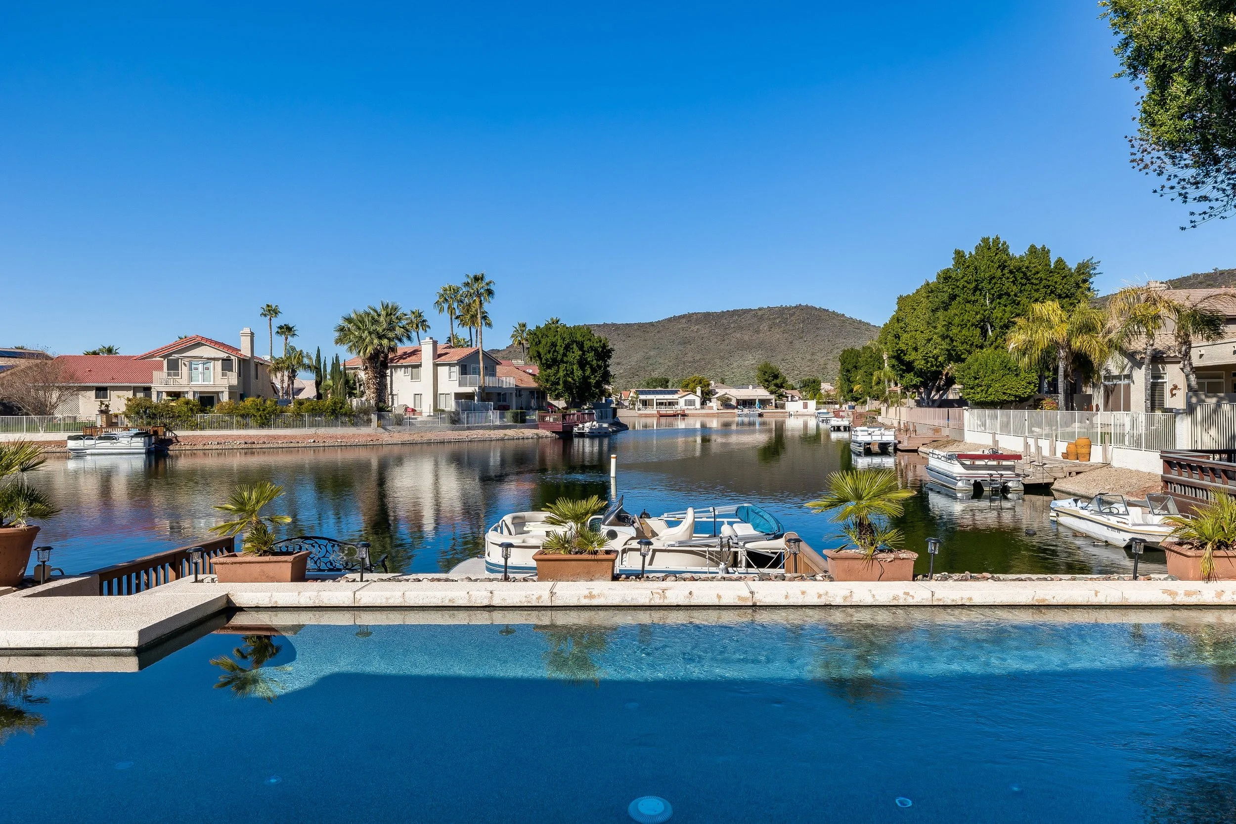 A canal lined with houses, boats, and palm trees under a clear blue sky, with a view from a swimming pool in the foreground.