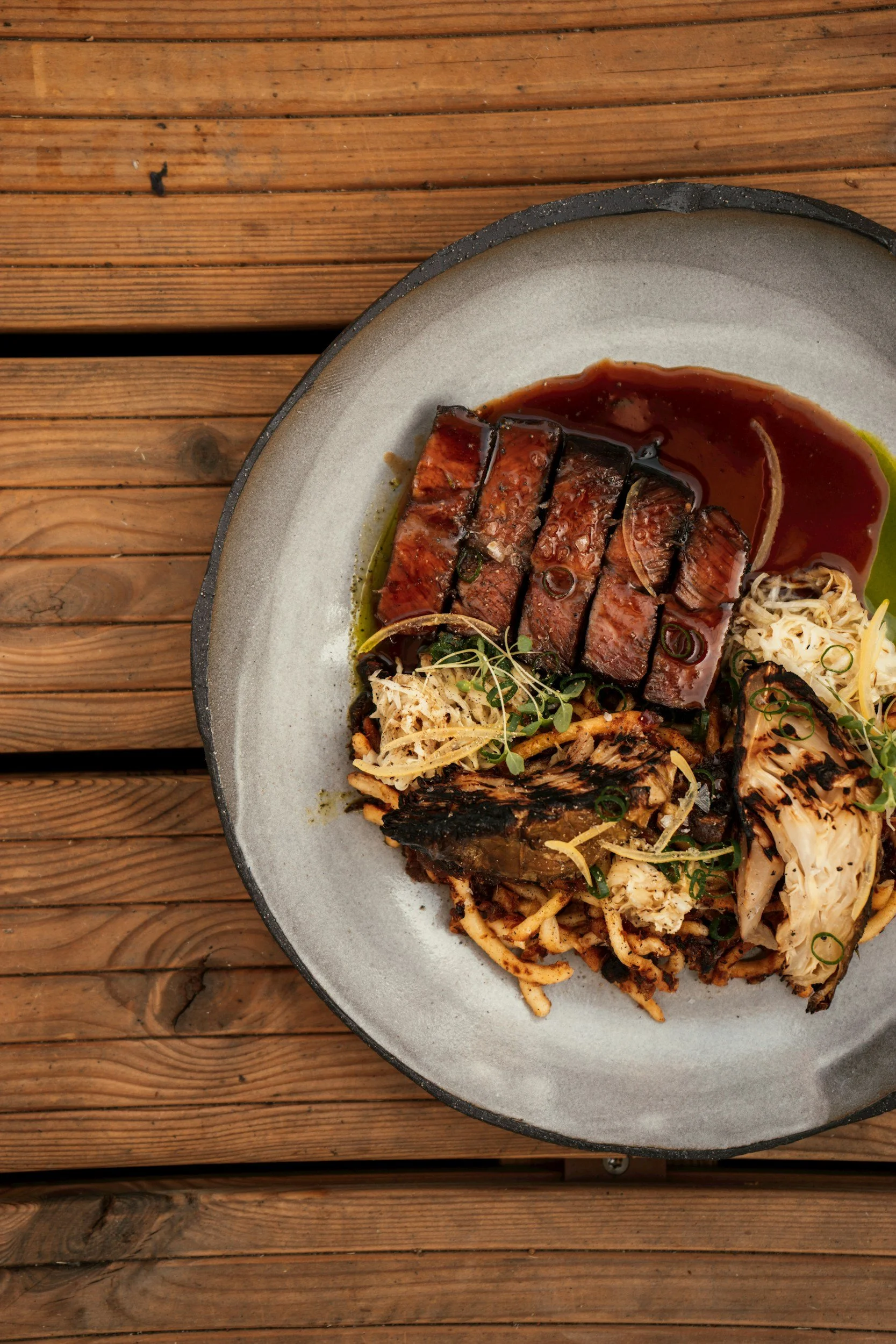 Close-up of a plate with sliced glazed meat, noodles, and vegetables on a wooden surface.