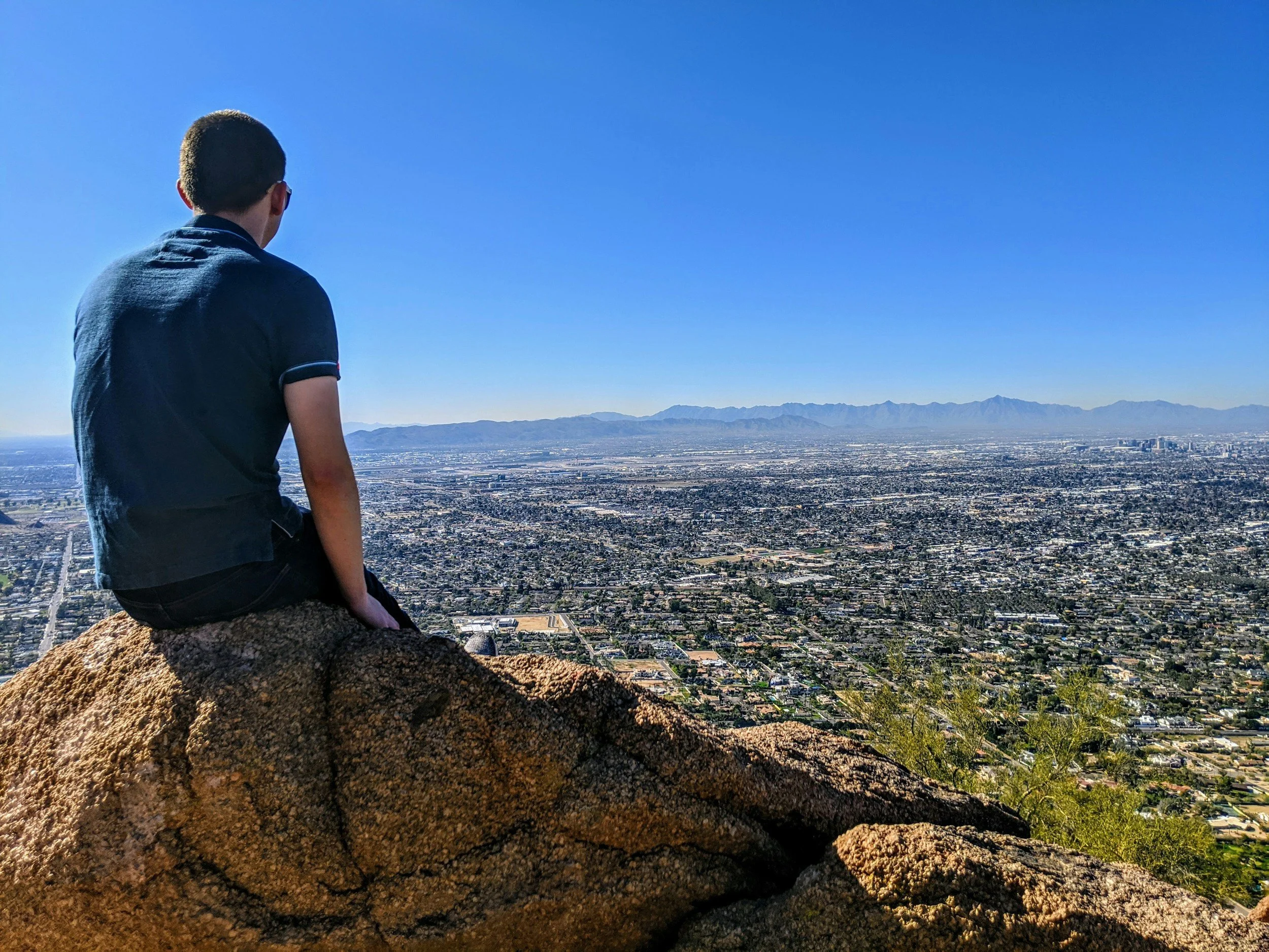 A person sitting on a rock, overlooking a cityscape with mountains in the distance on a clear day.
