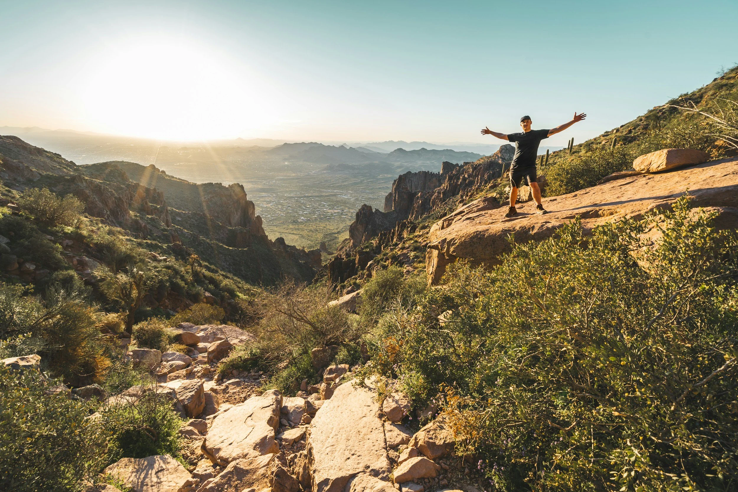 A person standing on a rocky mountain ledge with arms outstretched, overlooking a desert valley with mountains in the distance during sunrise or sunset.