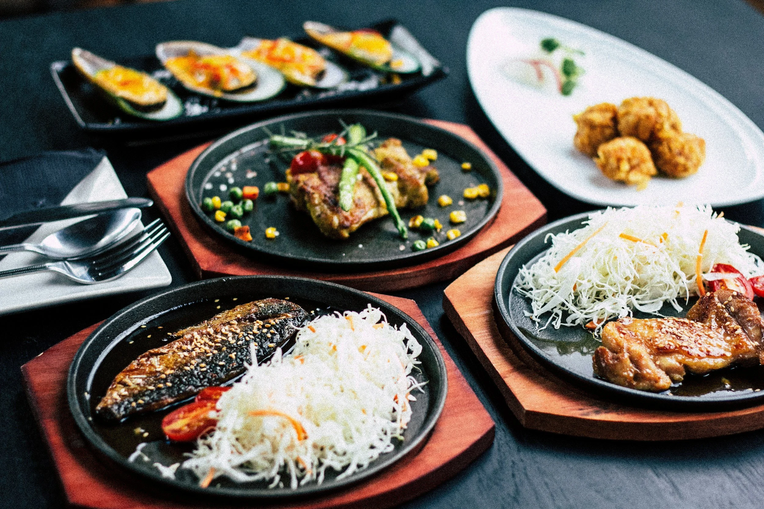 Variety of Asian dishes served on black and wooden plates, including grilled fish, fried chicken, stuffed mussels, shredded cabbage, and mixed vegetables, all arranged on a dark table.