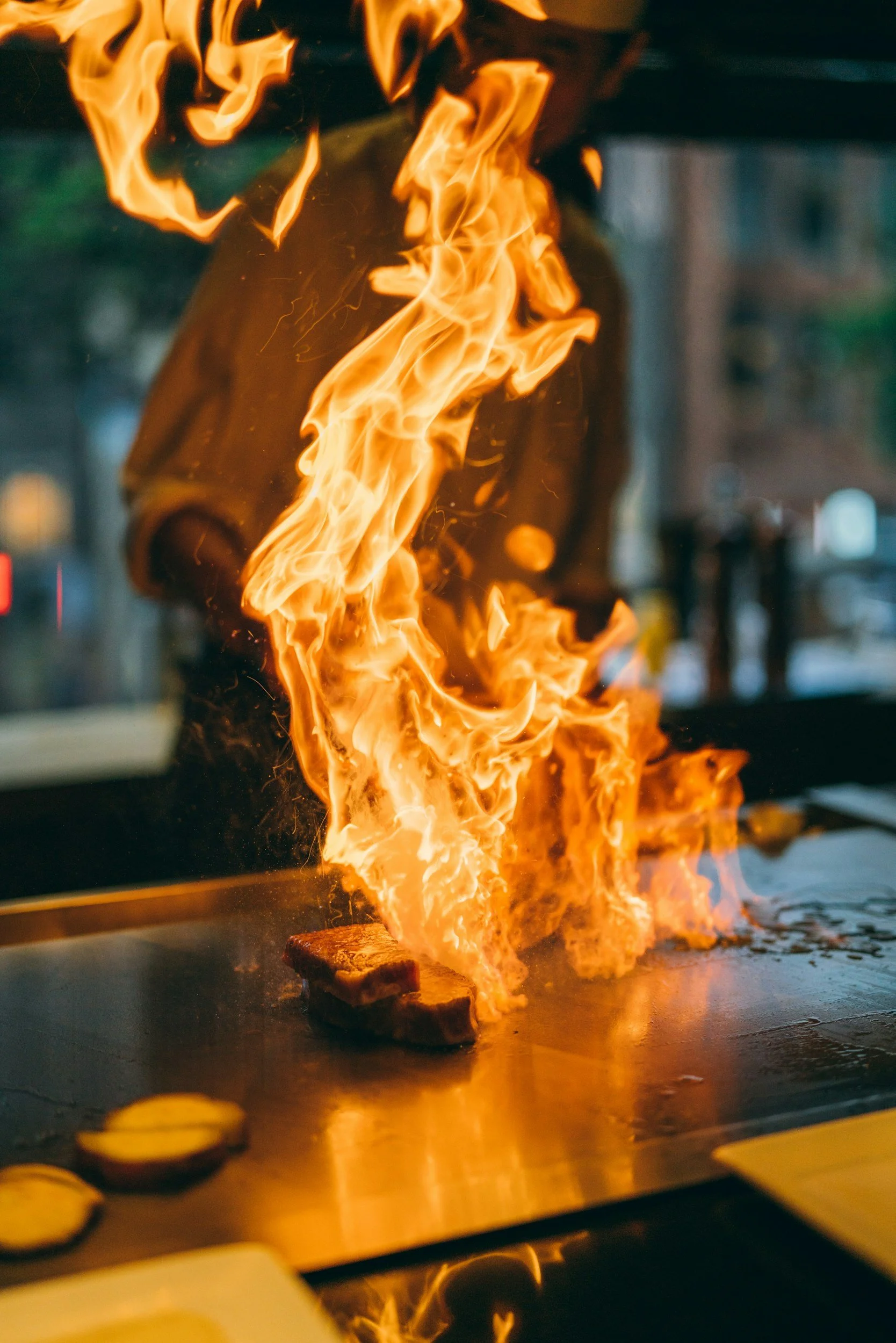 Flames and fire engulfing a piece of meat on a grill or flat-top surface, with some sliced vegetables in the foreground.