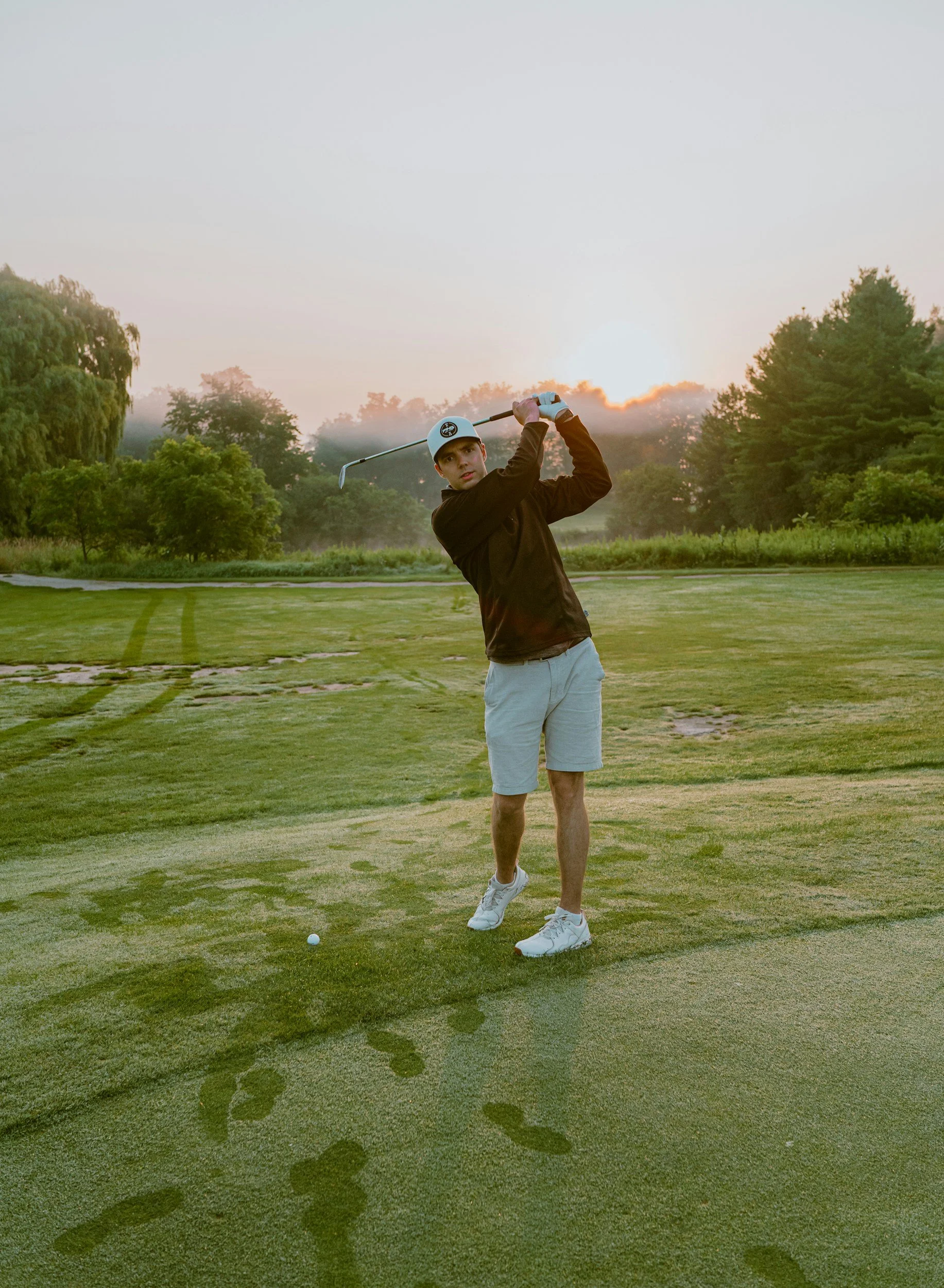 A young man in a black jacket, white shorts, and white golf shoes swings a golf club on a golf course at sunrise or sunset, with trees and a foggy sky in the background, leaving footprints in the grass.