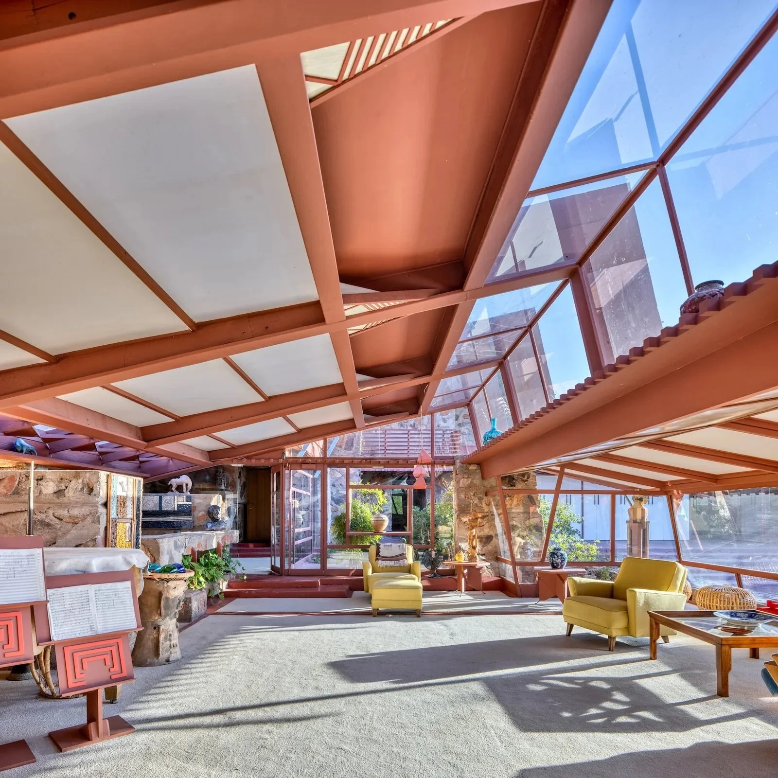 Interior view of a living space with large glass windows, brown wooden beams, and modern furniture including yellow armchairs, a coffee table, and decorations. Sunlight streams into the room.