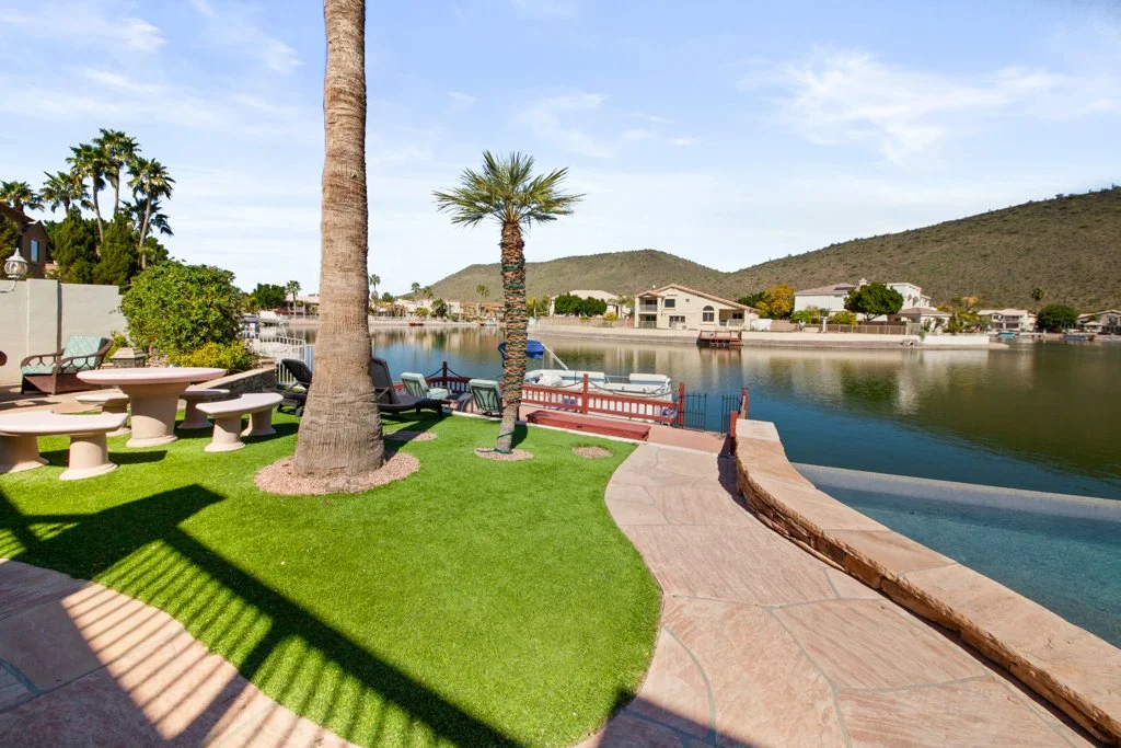 A backyard with a grassy area, palm trees, stone patio, seating, and a view of a lake with houses and hills in the background.