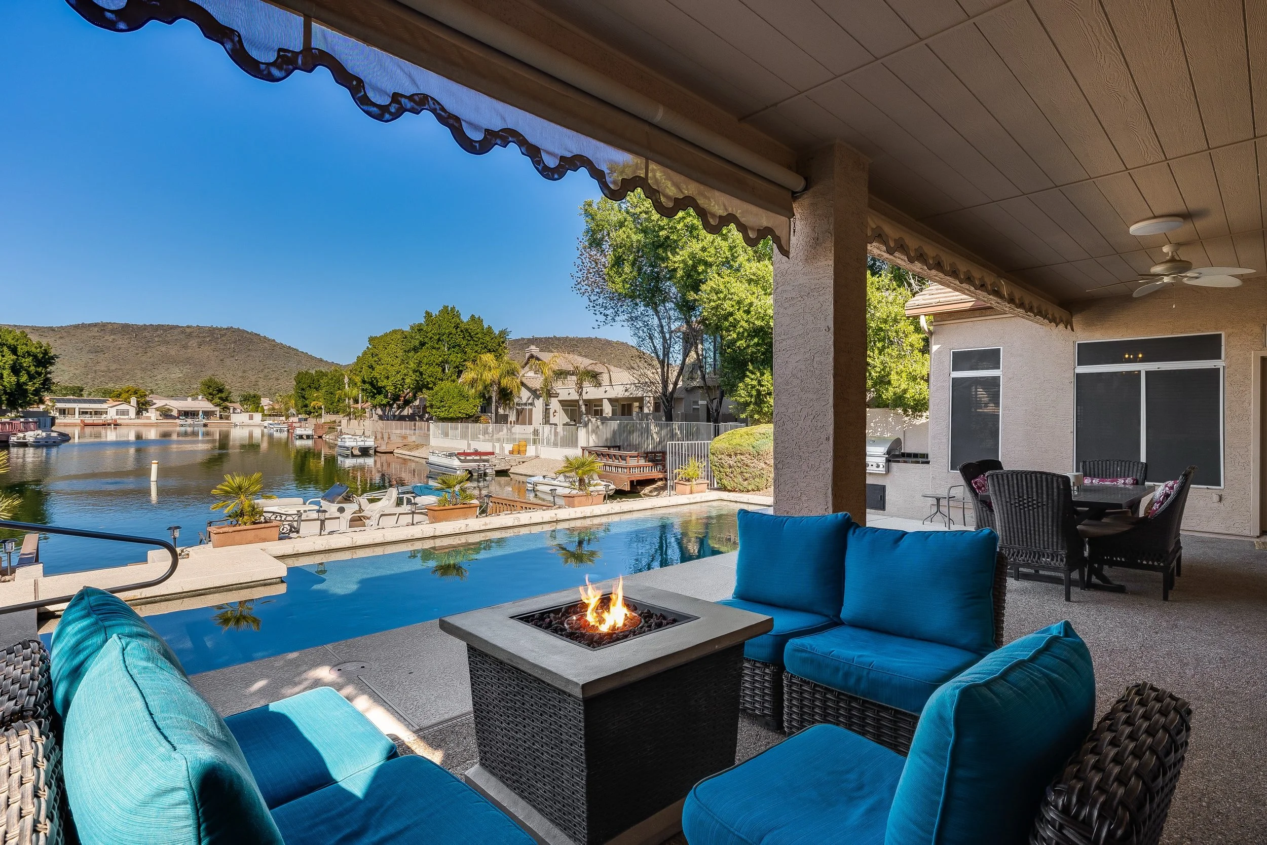 Covered patio overlooking a backyard with a pool, sitting area with blue and black outdoor furniture, a fire pit, and a view of the canal with boats and houses, hills in the background
