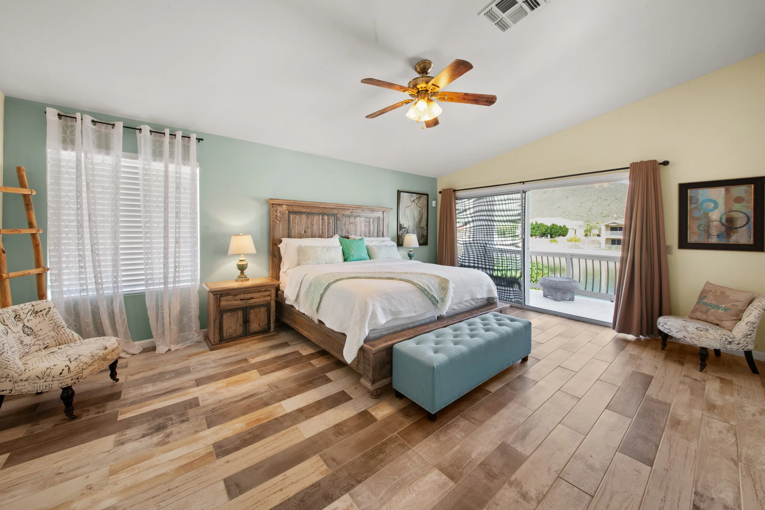 A bedroom with a wooden bed frame, white bedding, and green pillows. There are two nightstands with lamps, a patterned chair, and a sliding glass door with curtains. The room has light-colored walls and wood plank flooring.