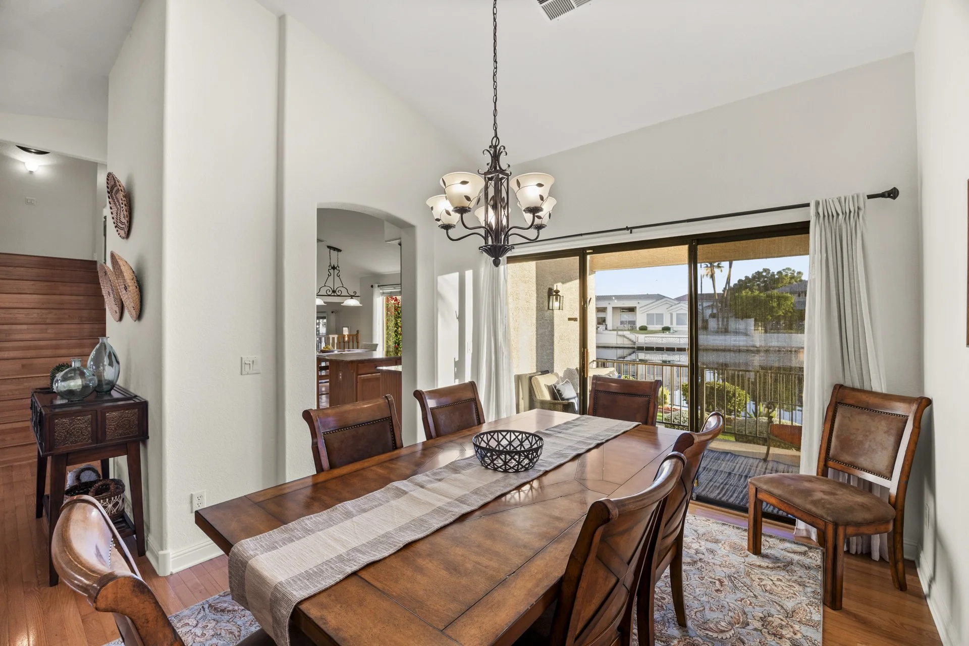 A dining room with a wooden table, six matching chairs, and a decorative runner. A sideboard with glass vases is on the left, and a sliding glass door with curtains leads to an outdoor balcony with view of water and houses. There is a chandelier hang
