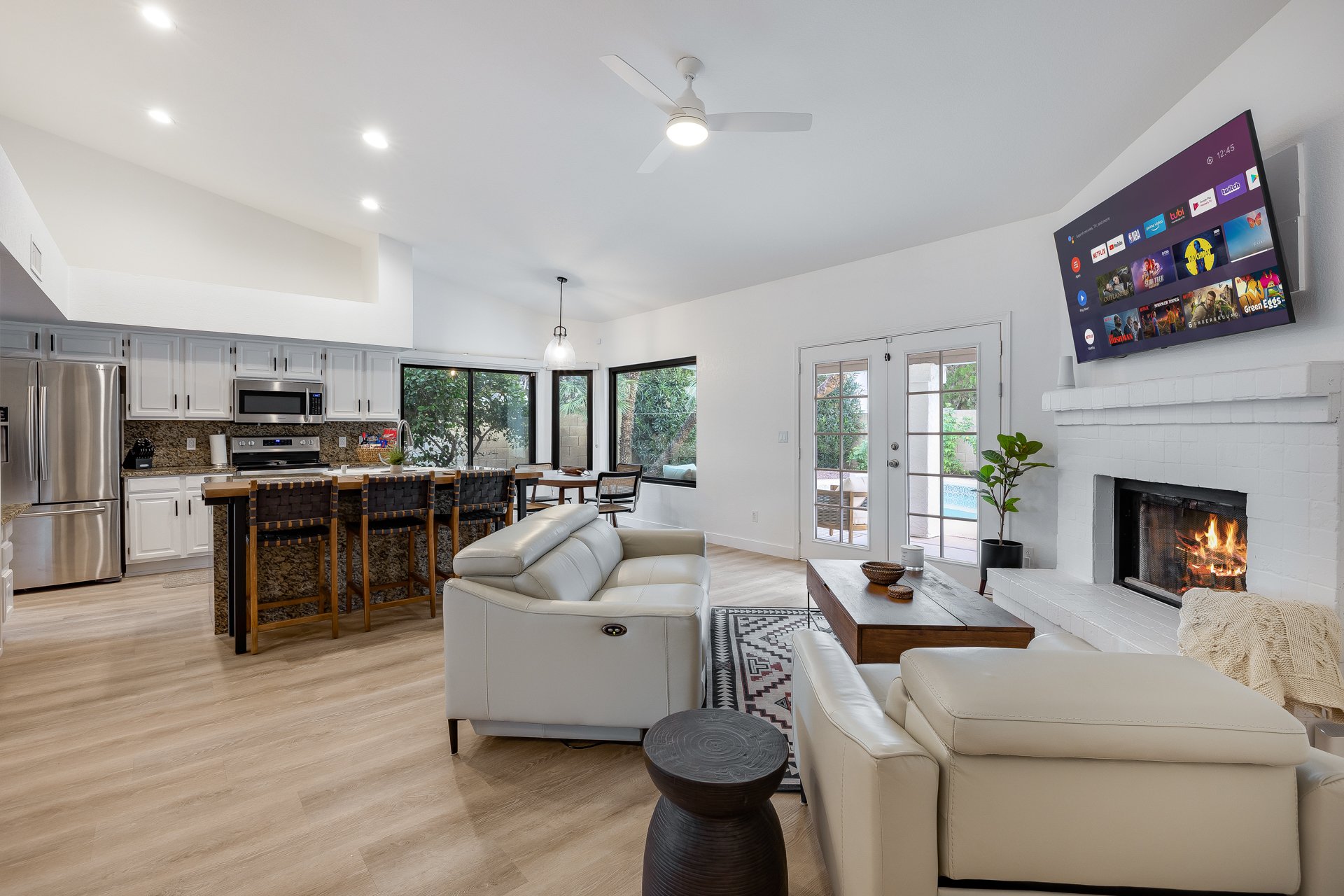 Open-concept living room and kitchen with white walls, wood flooring, ceiling fan, and large windows showing greenery outside. Features include a cozy fireplace, mounted TV, and modern furniture.