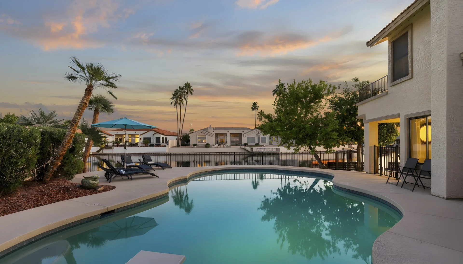 Residential swimming pool area with lounge chairs, umbrella, and surrounding greenery at sunset, with neighboring houses in the background.