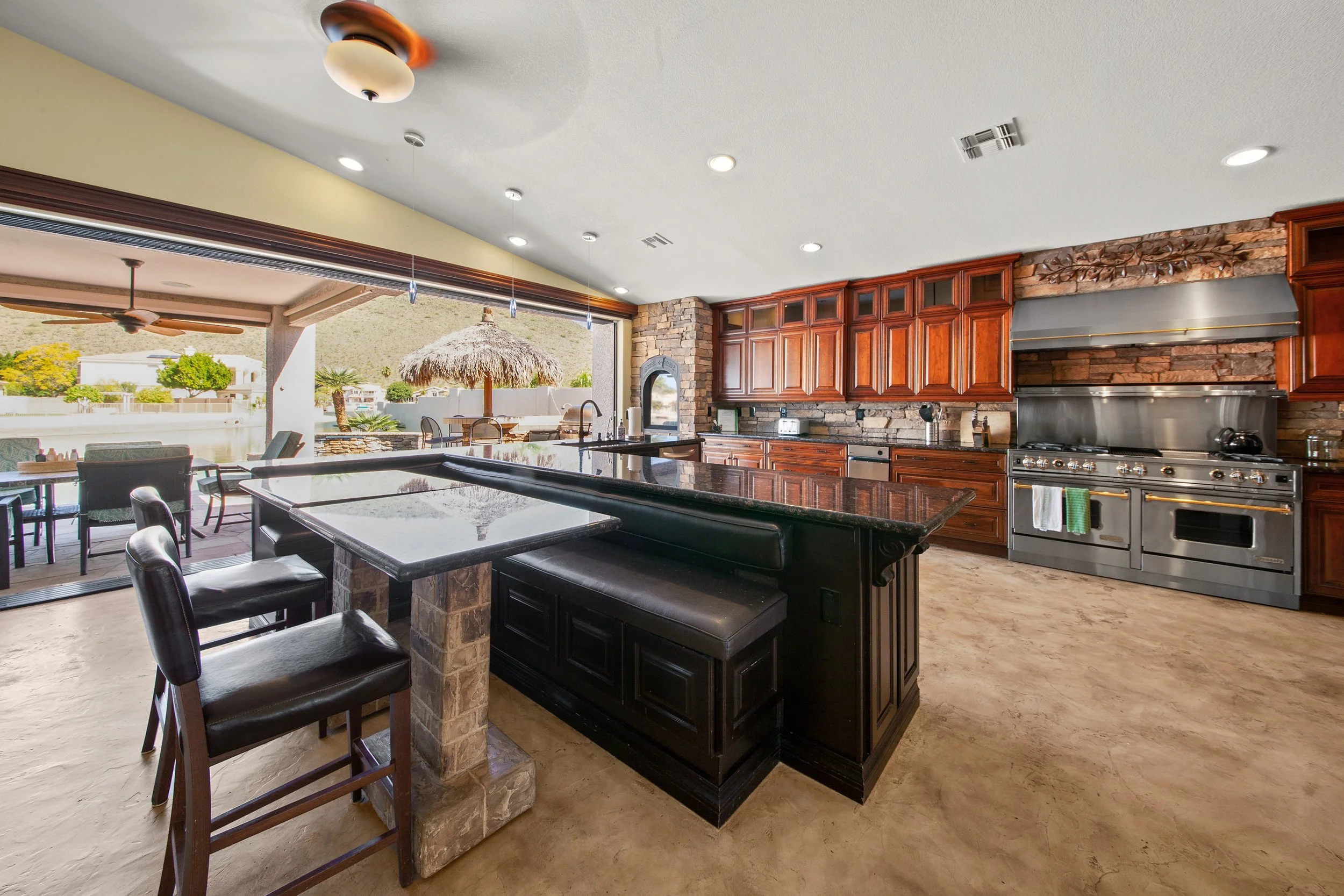 Open kitchen with wooden cabinets, stainless steel stove, black kitchen island with barstools, and outdoor patio with seating and a thatched umbrella, scenic mountain view.
