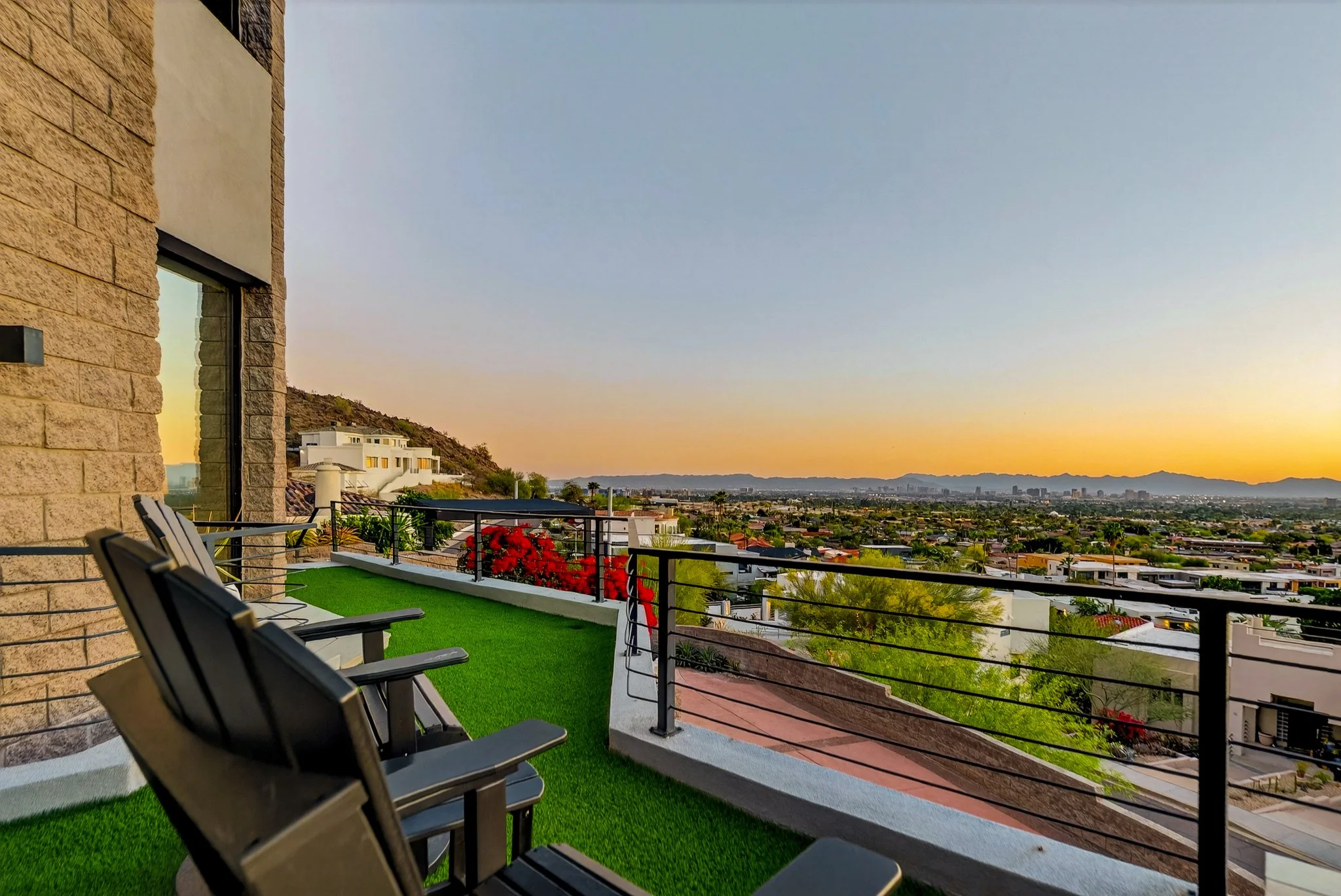 Modern balcony at sunset with black chairs, green artificial grass, red flowers in a planter, and cityscape in the distance under a colorful sky.