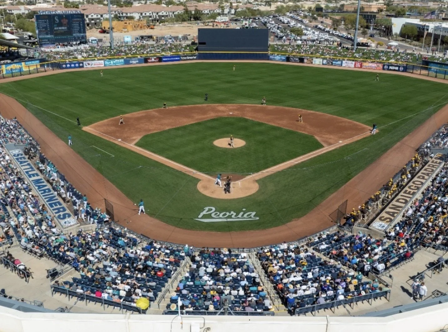 Aerial view of a baseball stadium filled with spectators, with players on the field, and advertising banners along the outfield fence that read 'SEATTLE MARINERS' and 'SAN DIEGO PADRES'. The field has green grass and a dirt infield, with the word 'Peoria' painted near the home plate area.