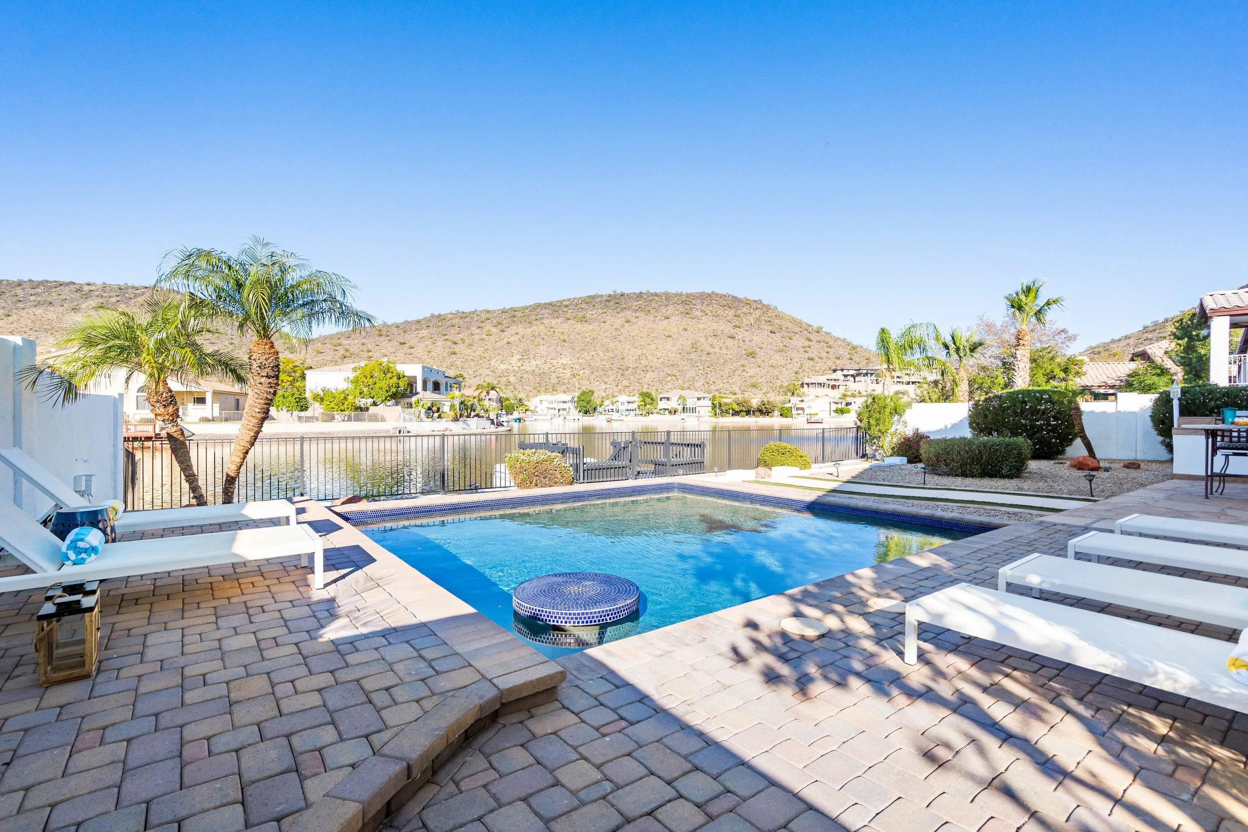Swimming pool area with lounge chairs, palm trees, and a view of houses and a mountain in the background under a clear blue sky.
