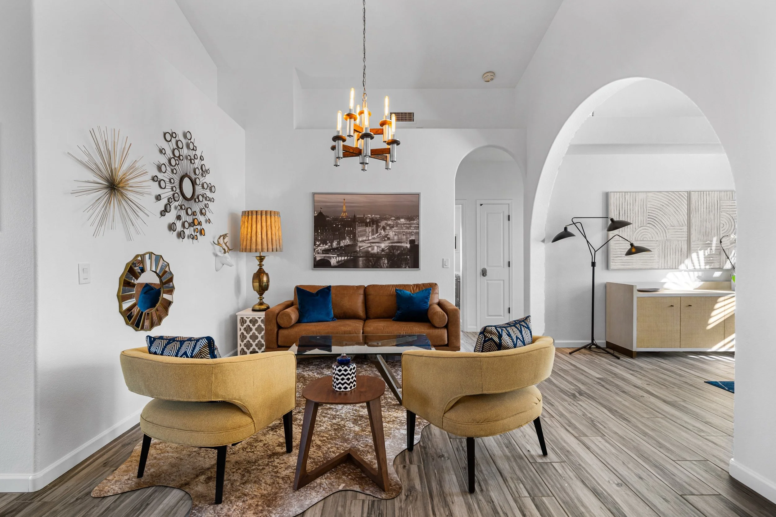 A living room with a brown leather sofa, yellow armchairs, a round glass coffee table, and decorative wall art. The room features a vintage-style floor lamp, a chandelier, and framed pictures on the walls.