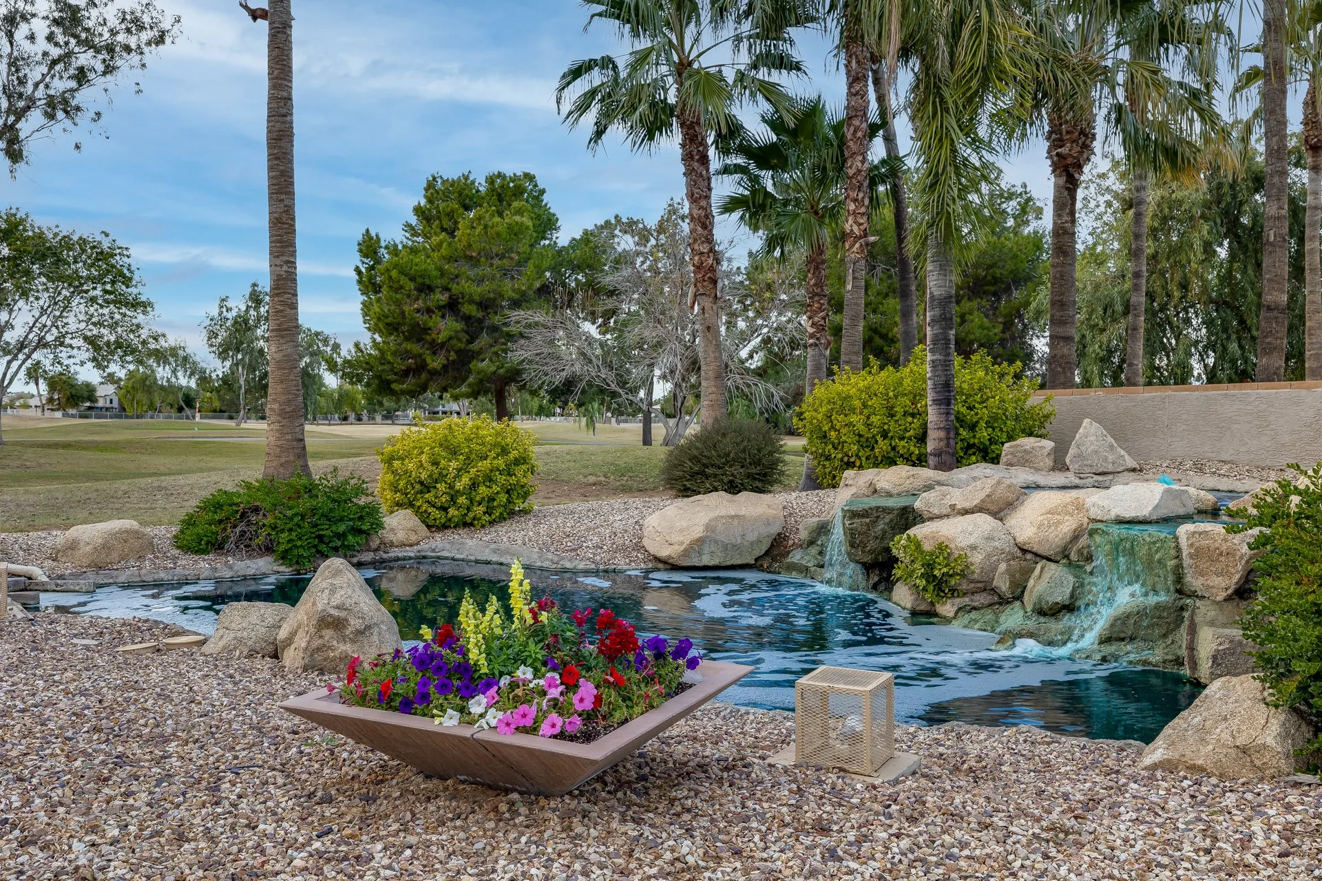 A backyard with a small pond featuring rocks and waterfalls, a flower bed with colorful flowers, and various palm trees and shrubs in the background.
