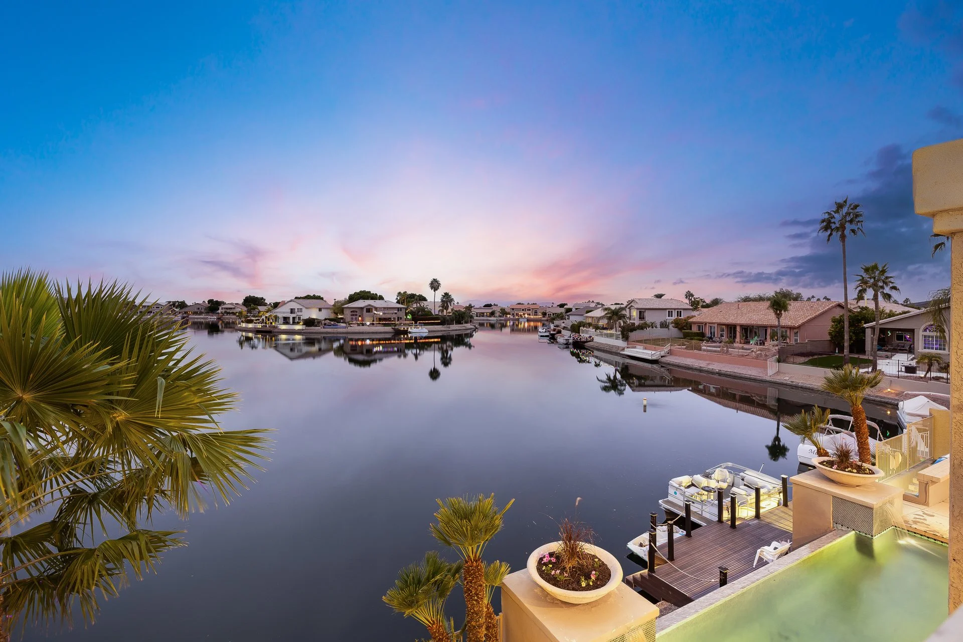 A serene waterfront scene during dusk or dawn with calm water reflecting houses, palm trees, and a colorful sky with pink and purple hues.