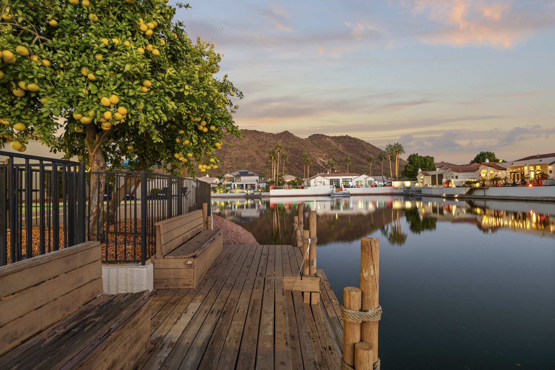 A lakeside scene at dusk with a lemon tree in the foreground, calm water reflecting homes and palm trees, mountains in the background, and a colorful sky.
