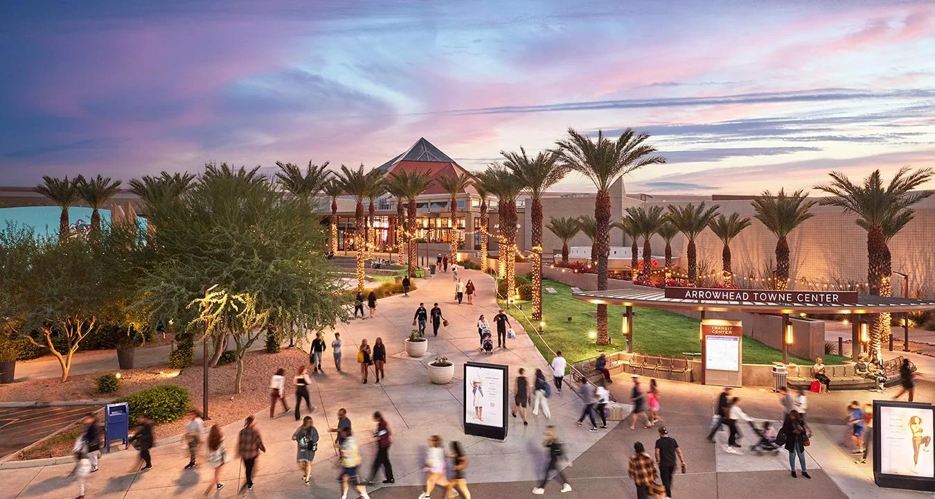 People walking outside Arrowhead Towne Center shopping mall during sunset, with palm trees, lit pathway, and shopping mall in the background.