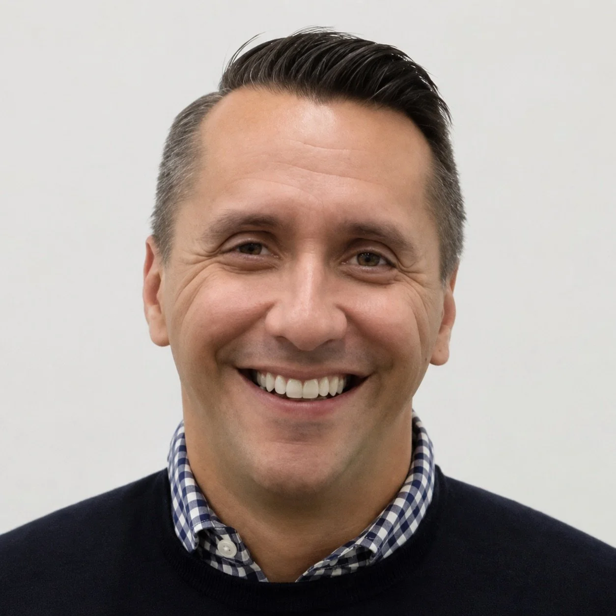 A man with short dark hair, smiling, wearing a dark sweater over a checkered shirt, standing against a plain light background.