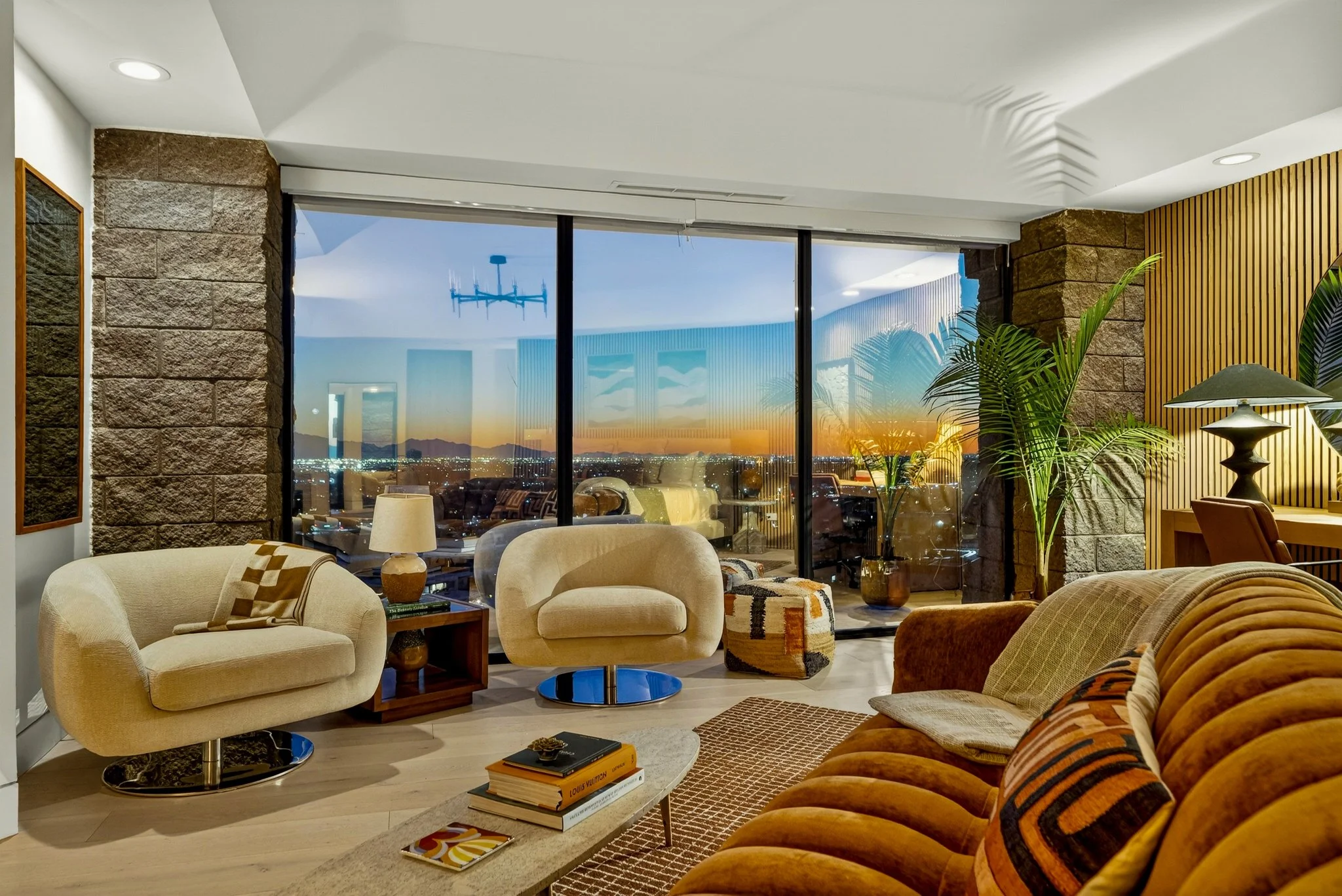Living room with beige and brown upholstered furniture, large windows showing city skyline sunset, and indoor plants.