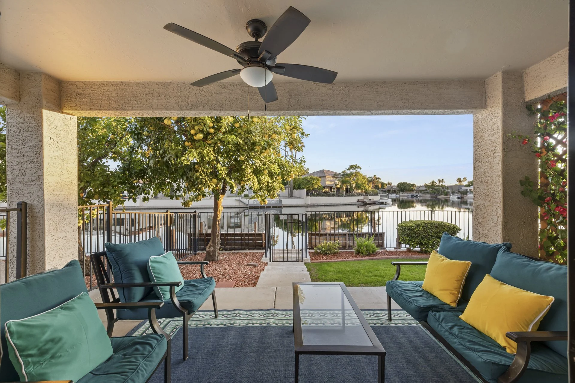 Covered patio with teal and yellow cushions, overlooking a backyard with a lemon tree, a waterway, and houses in the distance.