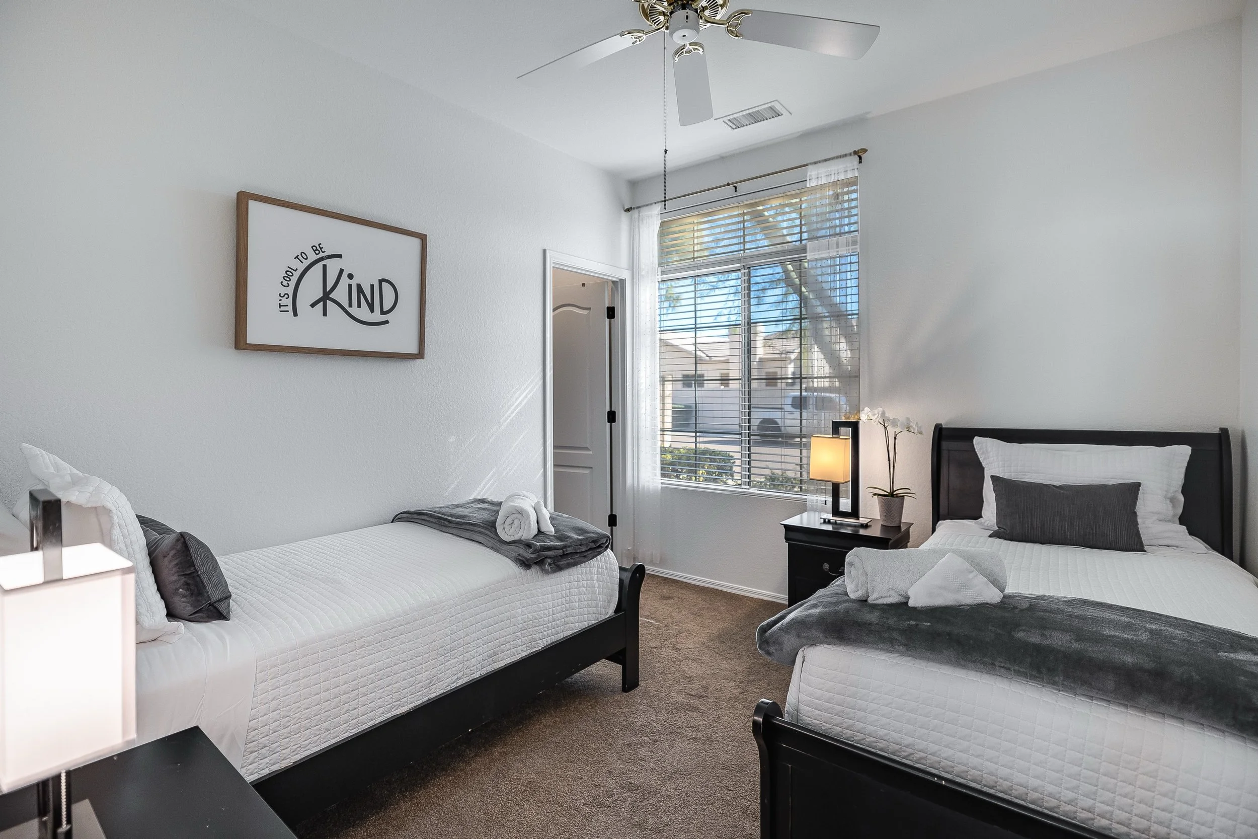 Two twin beds with white quilts and gray pillows in a bright bedroom with large window and a dresser with lamp and flowers.