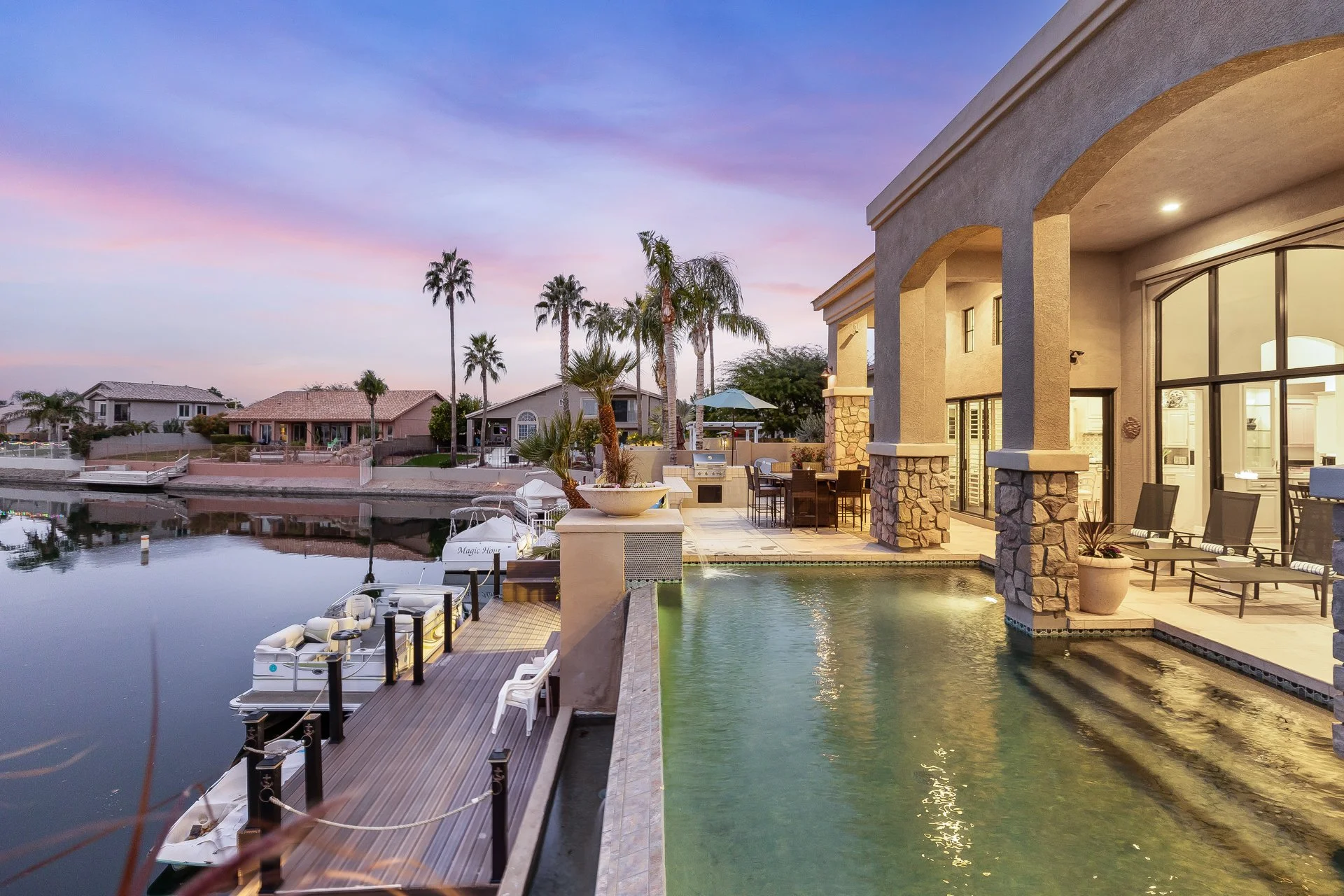 A backyard with a dock and boat on a calm waterway at sunset, featuring a house with large windows, outdoor furniture, and tall palm trees.