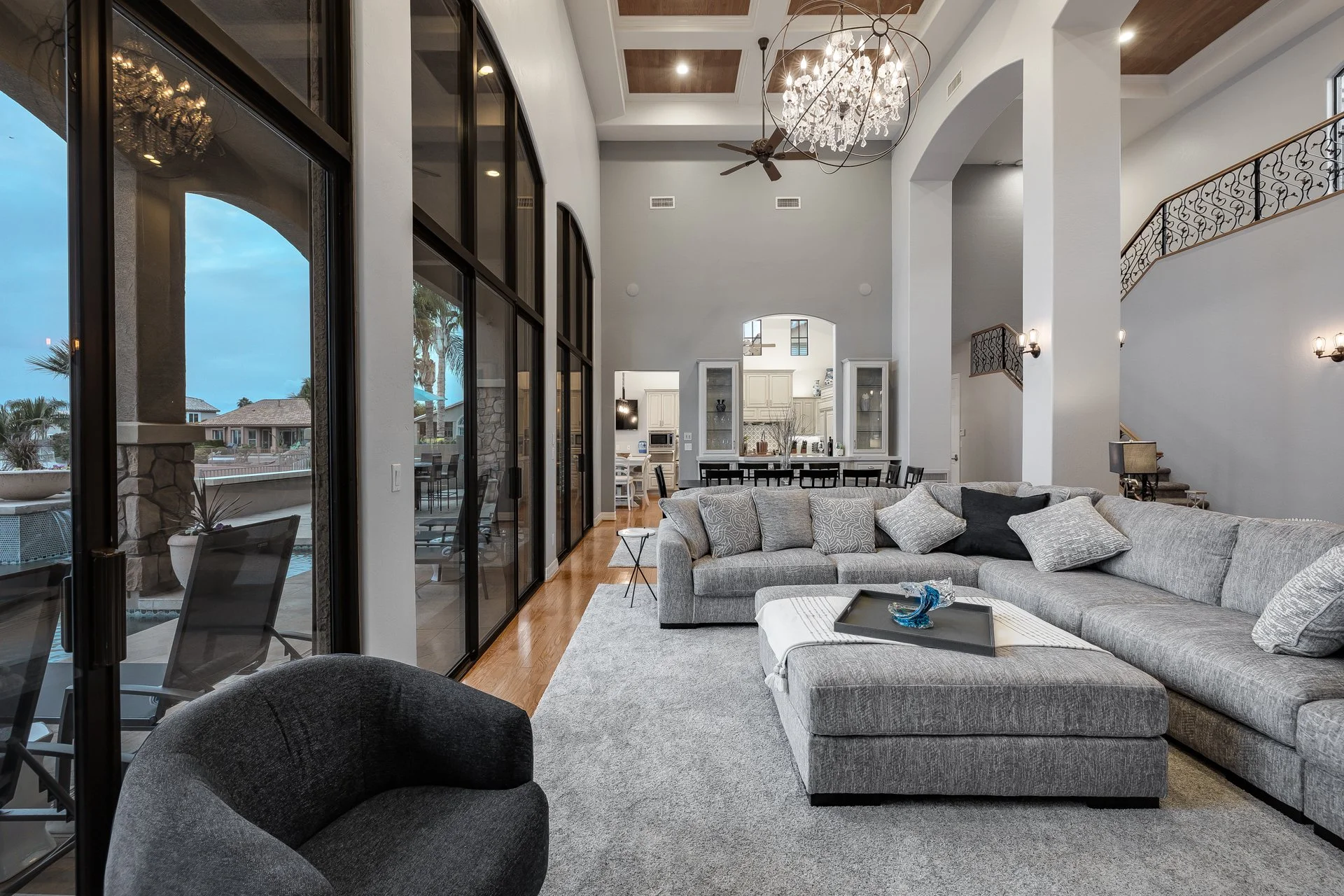 Spacious living room with large sectional gray sofa, black and gray armchair, wooden floor, and a chandelier hanging from a high ceiling. Glass sliding doors lead to an outdoor patio, with view of neighboring houses and palm trees. Open kitchen with 