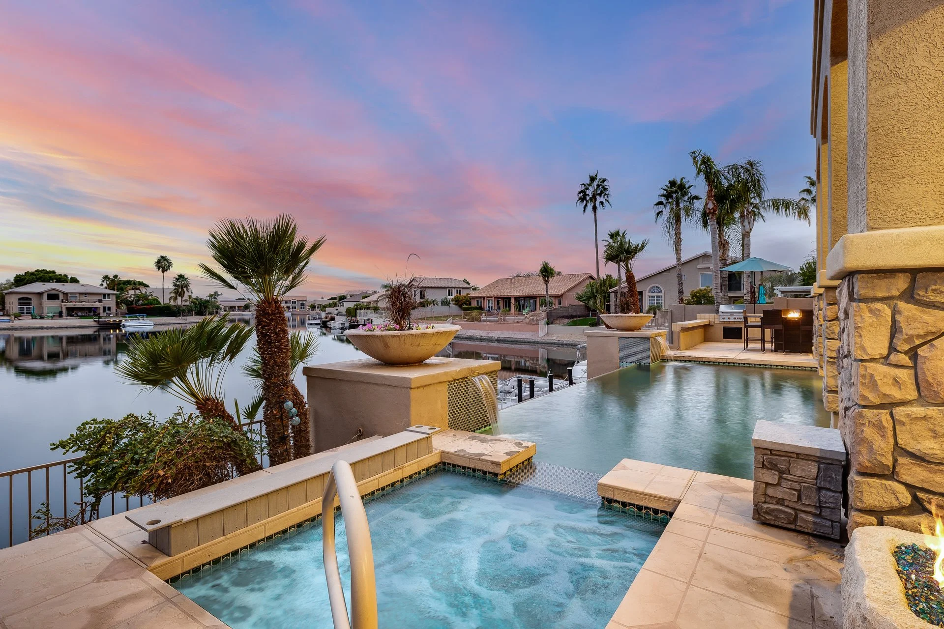 A backyard scene at sunset or sunrise with a swimming pool, palm trees, outdoor seating, a grill, and a canal with boats in a suburban neighborhood.