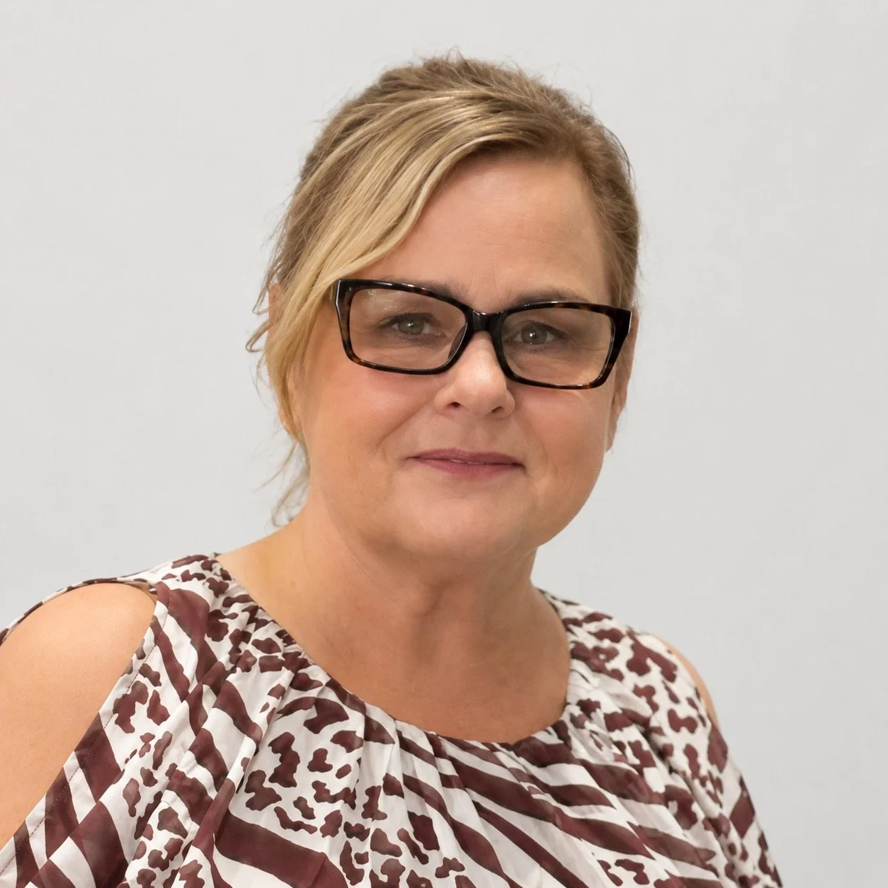 A woman with short blonde hair, wearing glasses and a patterned sleeveless top, standing against a plain light-colored background.