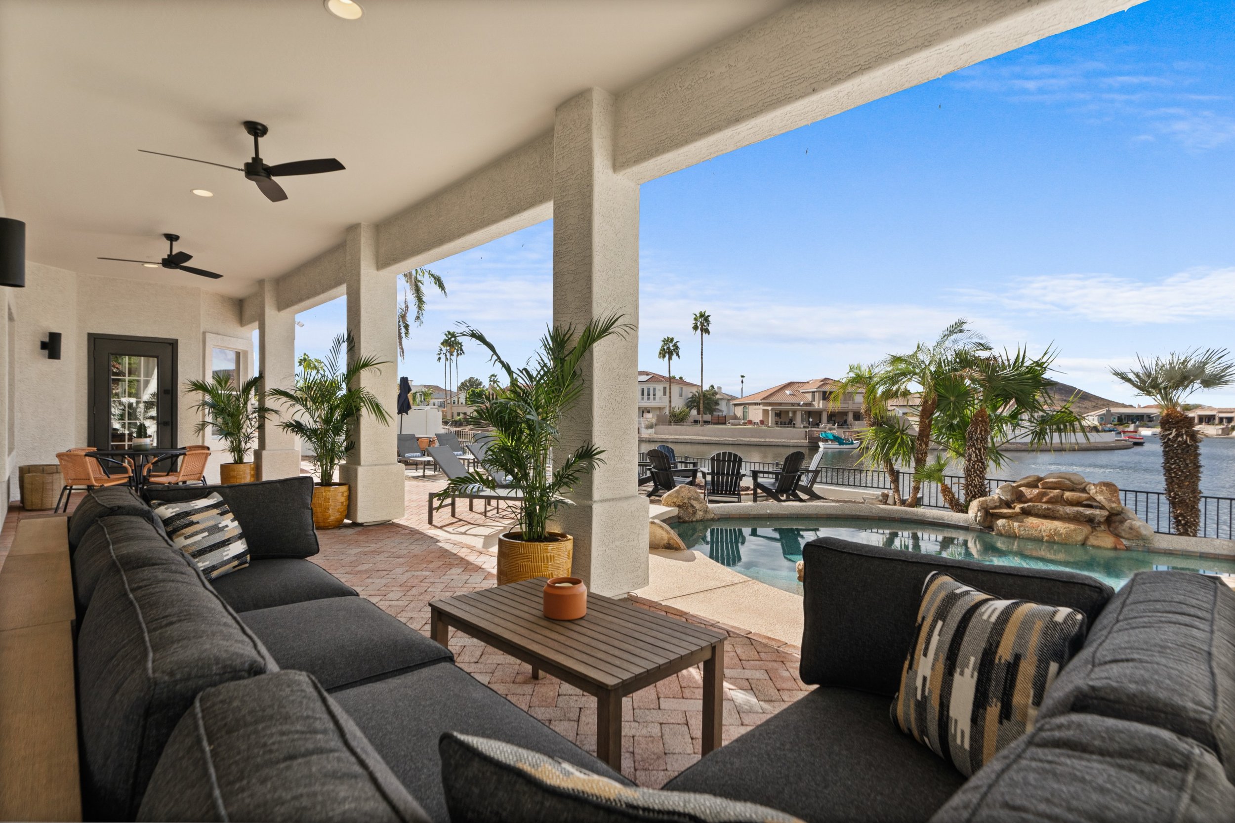 Covered outdoor patio with lounge seating, potted plants, and a view of a waterway with houses and palm trees.