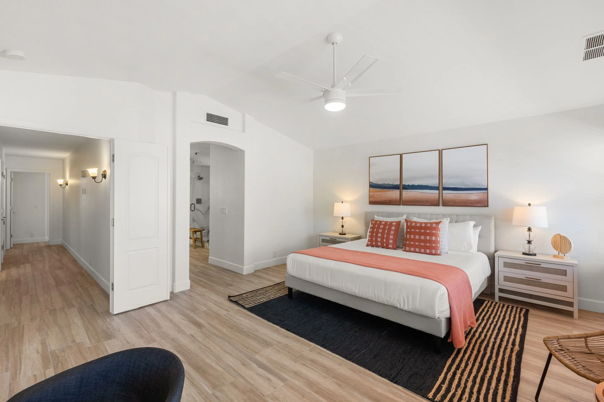 A bedroom with white walls and light wood flooring. The bed has white bedding, orange pillows, and a coral throw. There are two nightstands with lamps, and a triptych art piece of a beach scene above the bed. A ceiling fan is on the ceiling. Part of 