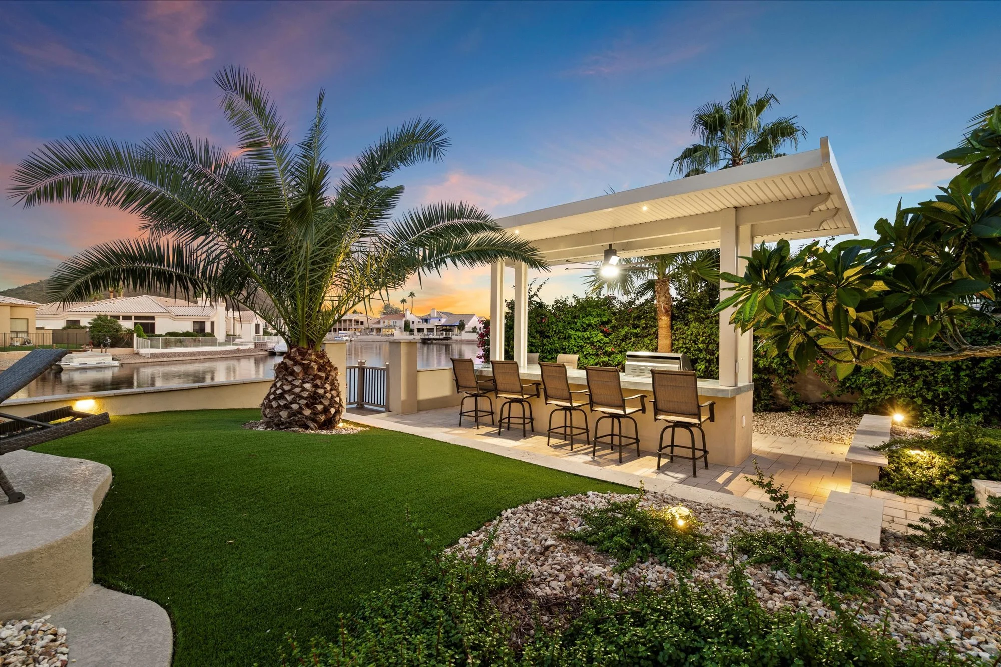 A backyard at sunset with a palm tree, green grass, a covered outdoor bar area with bar stools, and a waterfront view of houses and boats.