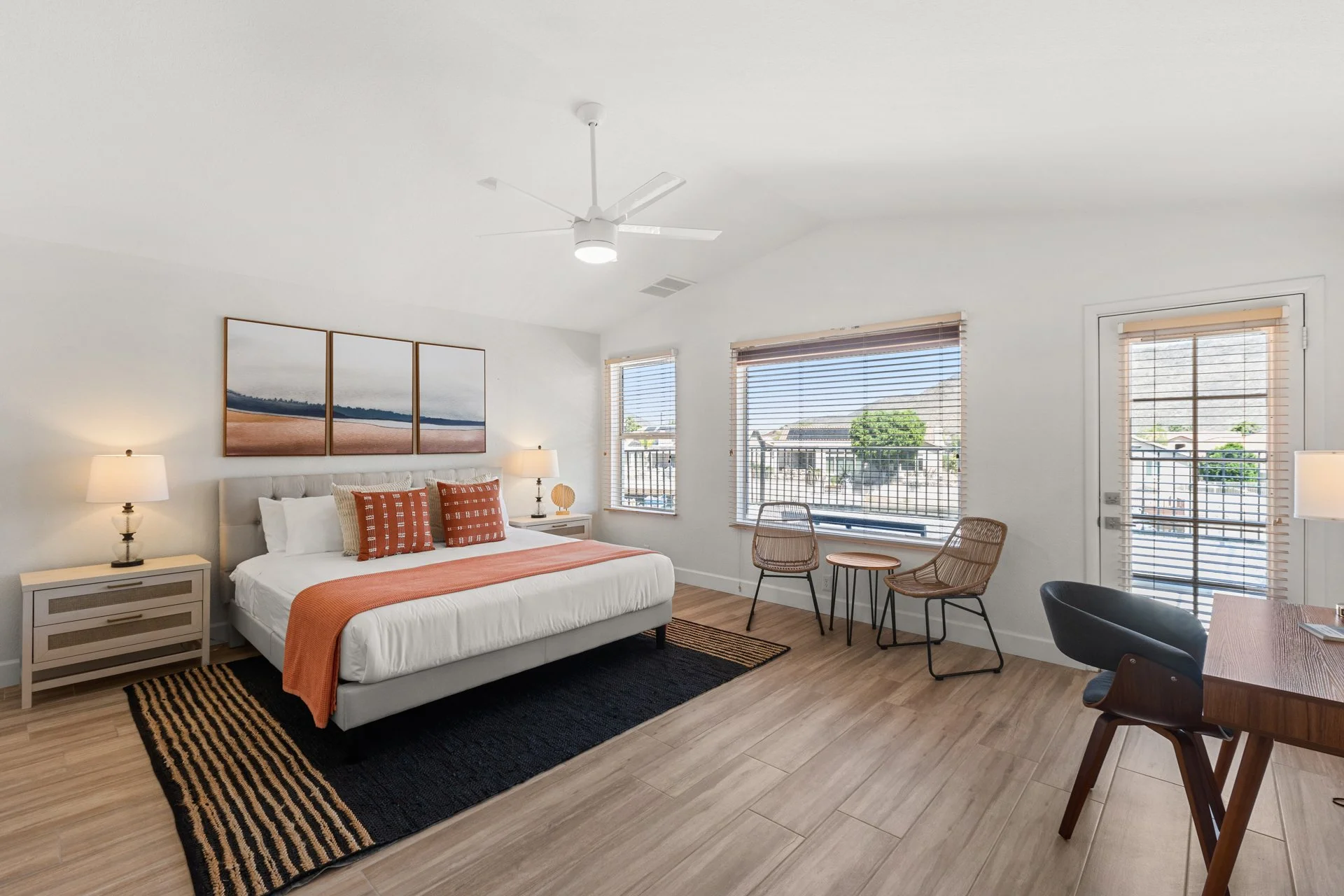 A modern bedroom with a white bed, orange accents, two nightstands with lamps, framed wall art, large windows with blinds, and a sitting area with rattan chairs and a door leading outside.