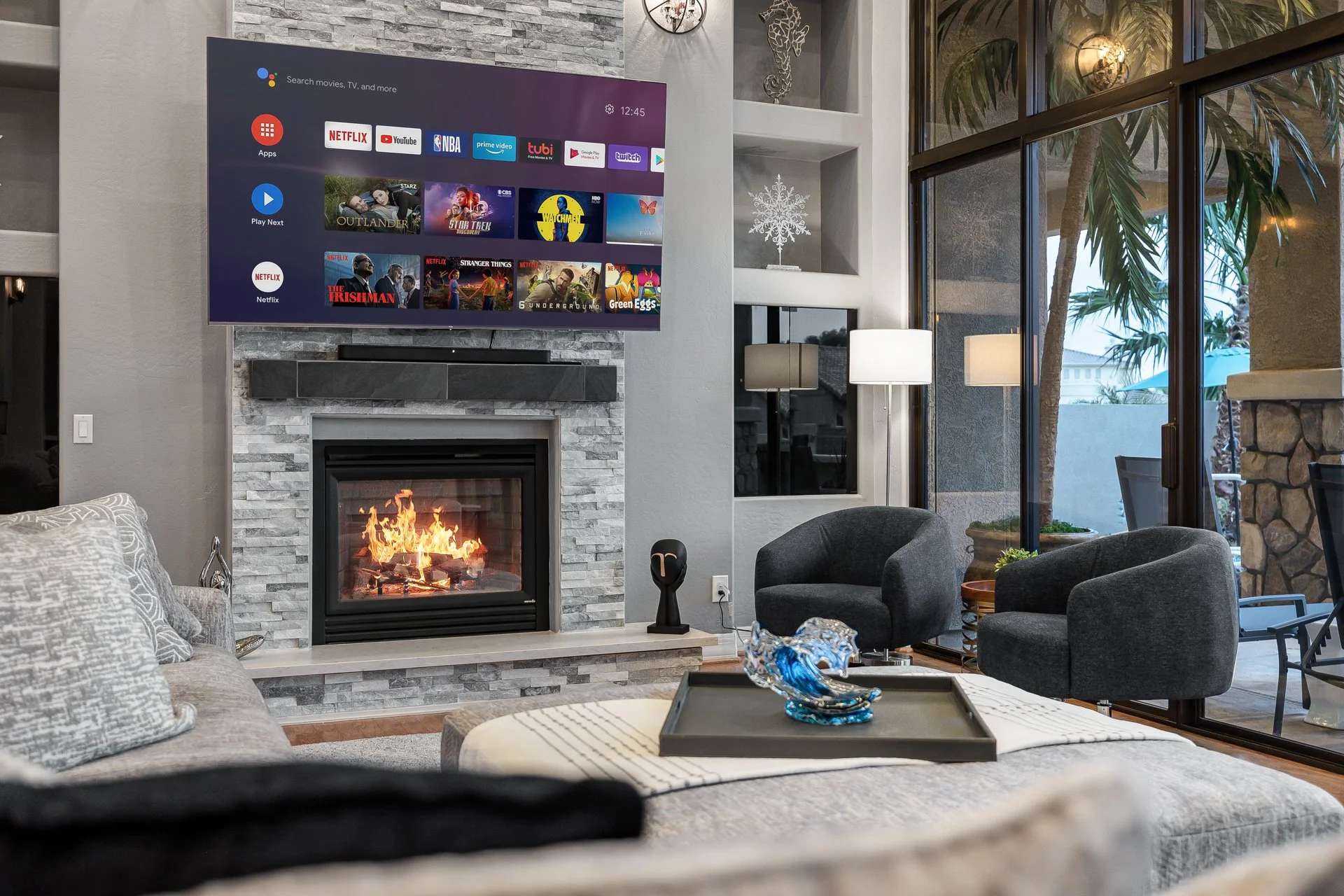 Living room with a flat-screen TV above a fireplace, modern gray furniture, and large windows showing outdoor plants and a patio.