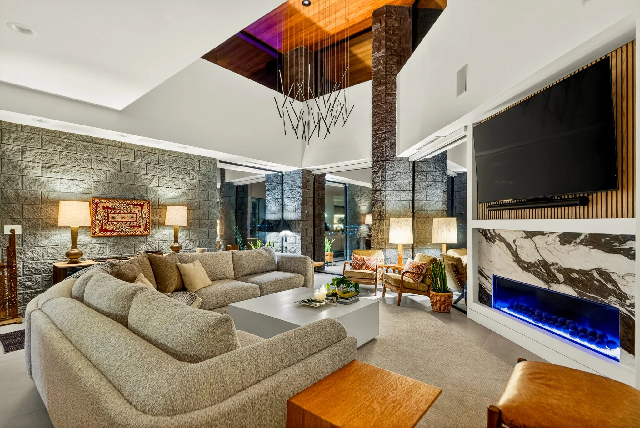 A spacious modern living room with a large beige sectional sofa, a white coffee table, and various chairs. The room has a stone accent wall, a fireplace with a black marble surround, and a large flat-screen TV mounted on a wooden slatted wall. There 