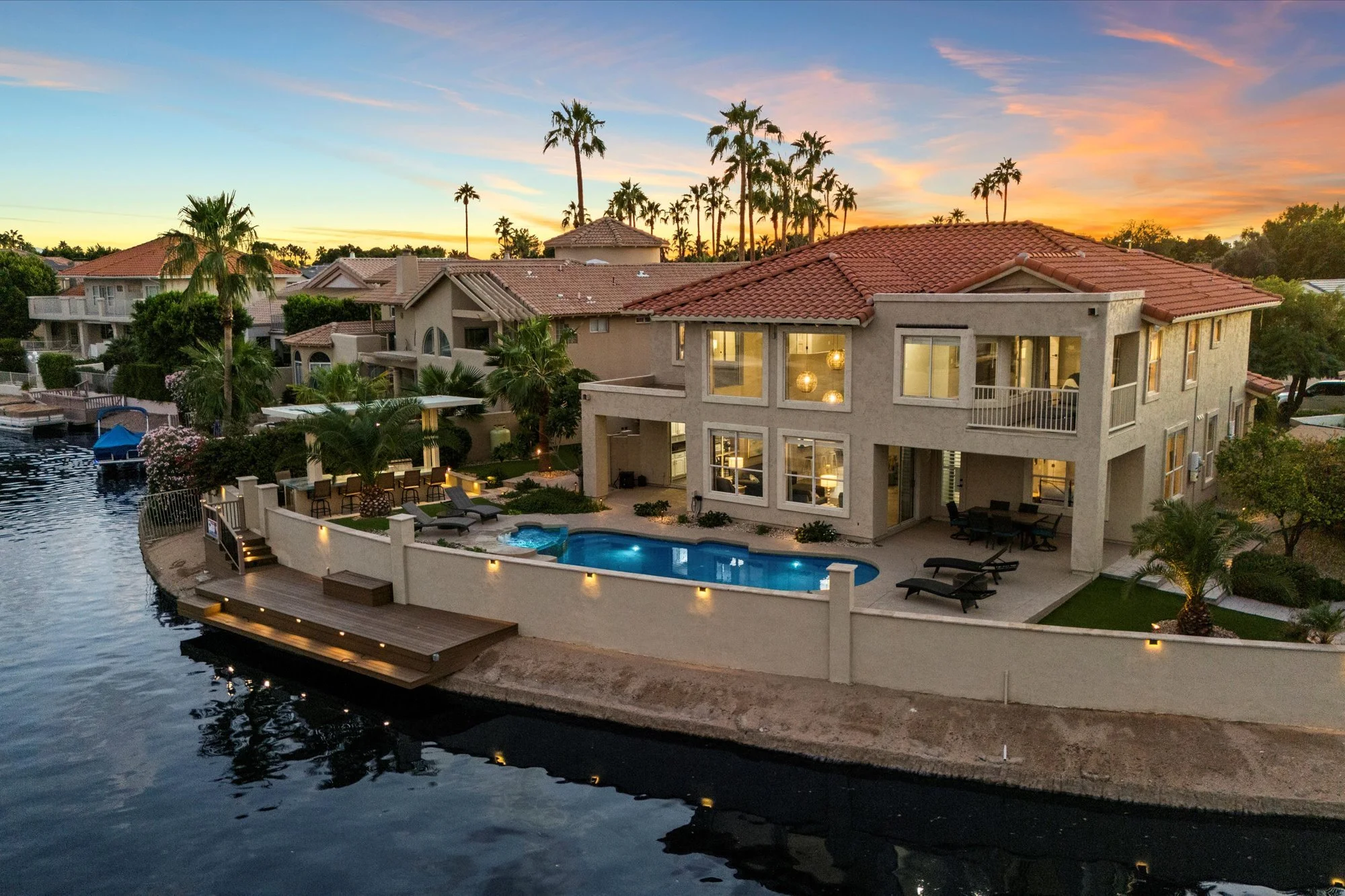 A luxurious lakeside house at sunset with large windows, an outdoor swimming pool, palm trees, and a dock extending into the water.
