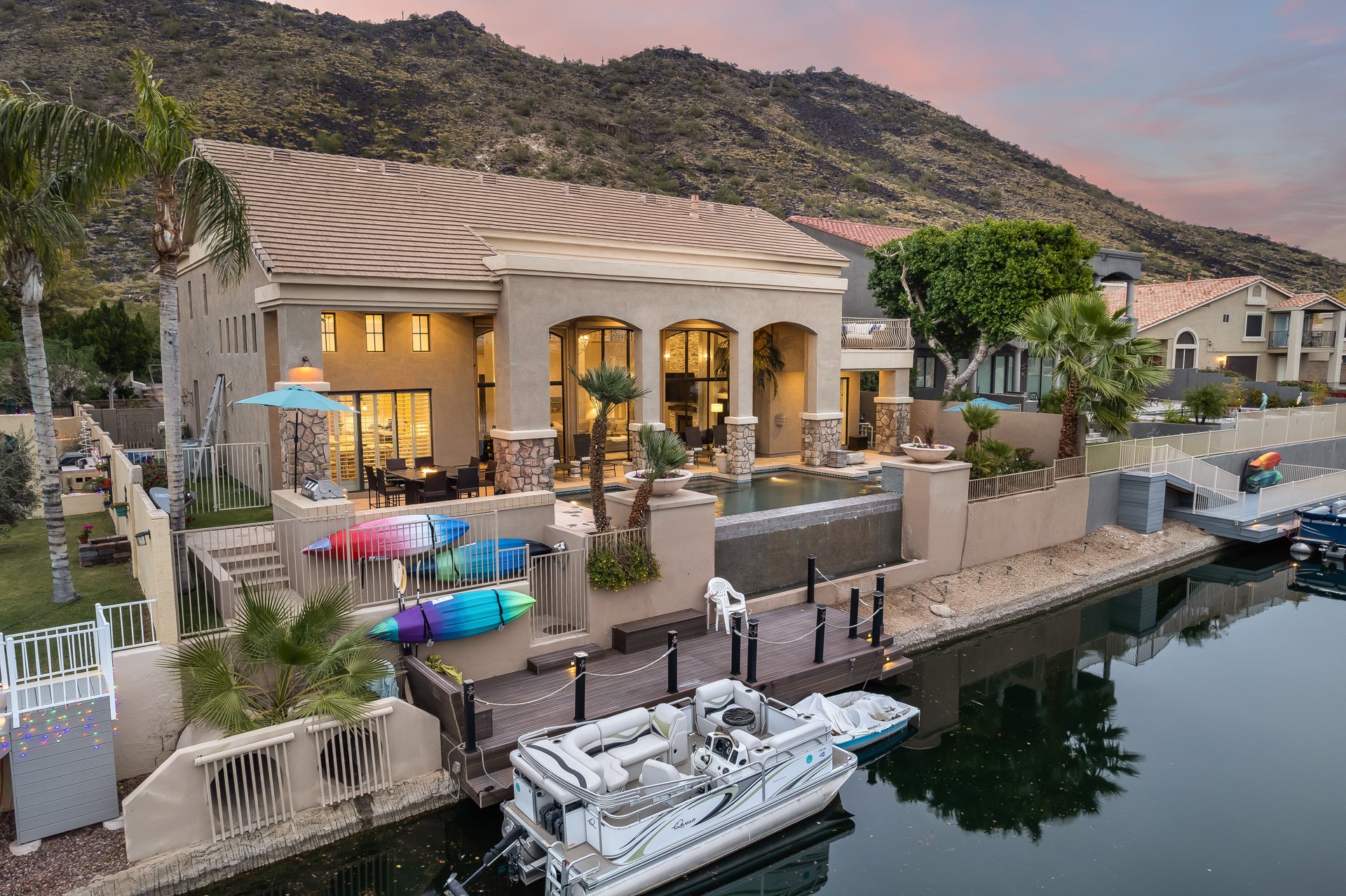 Modern house by a canal with boats, outdoor seating, palm trees, and kayaks, with mountains in the background during sunset.