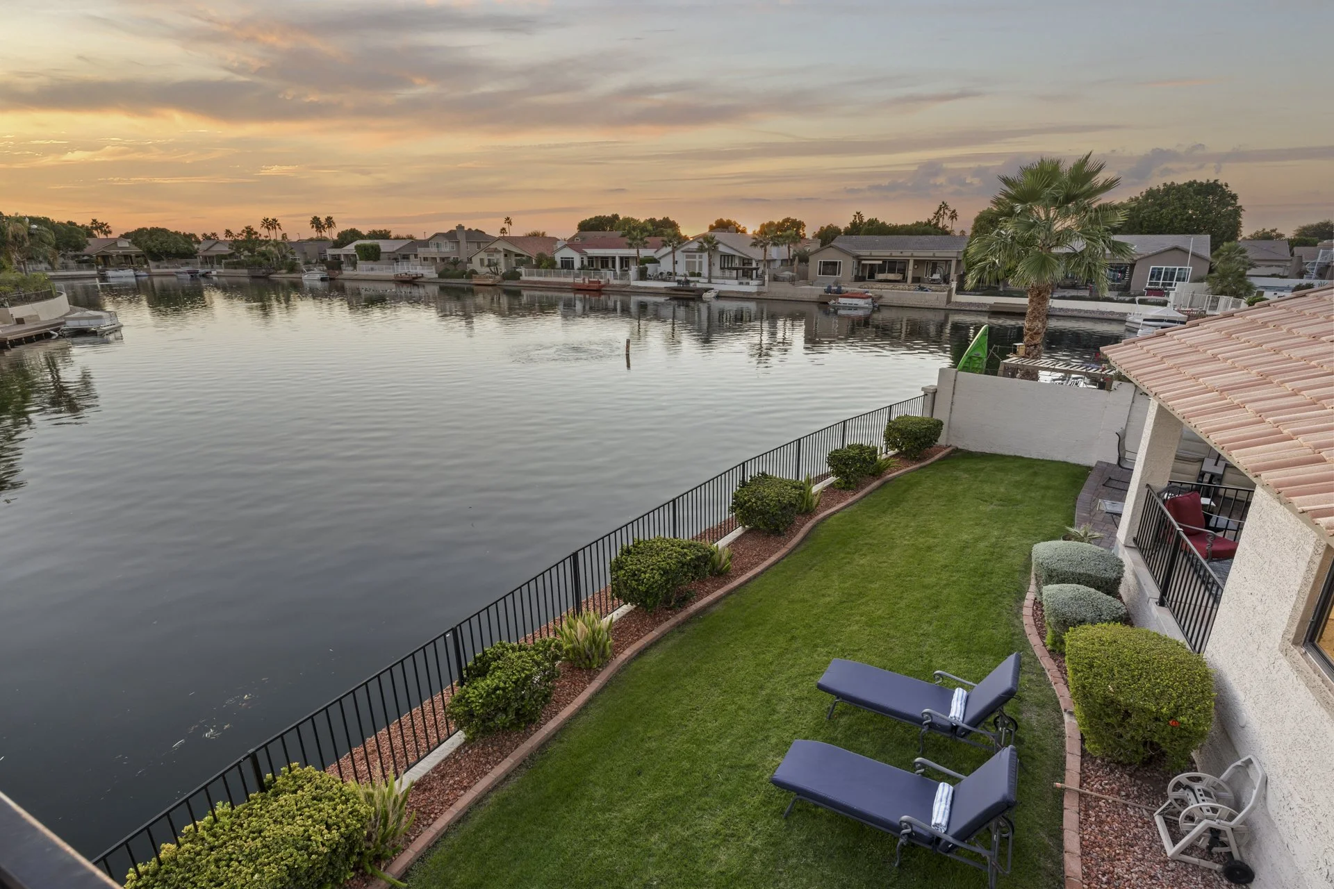 View of a calm water canal or lagoon with houses on the opposite side, during sunset with a colorful sky, seen from a backyard featuring green lawn, lounge chairs, a small patio, and a large palm tree.
