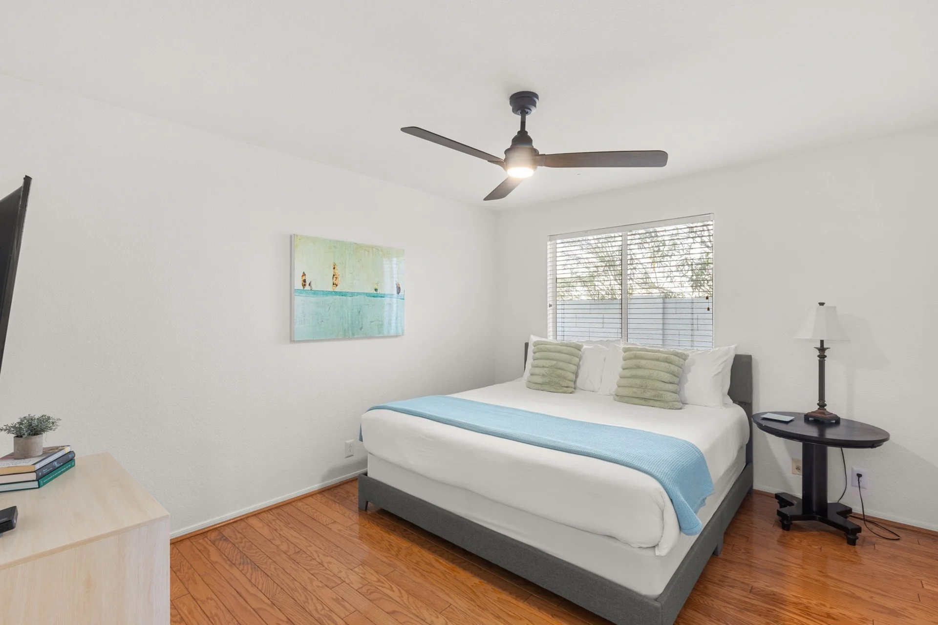 A bedroom with white walls and hardwood floors featuring a bed with white bedding and four pillows, a round black side table with a lamp, a wall painting of a beach scene, a window with blinds, and a ceiling fan.