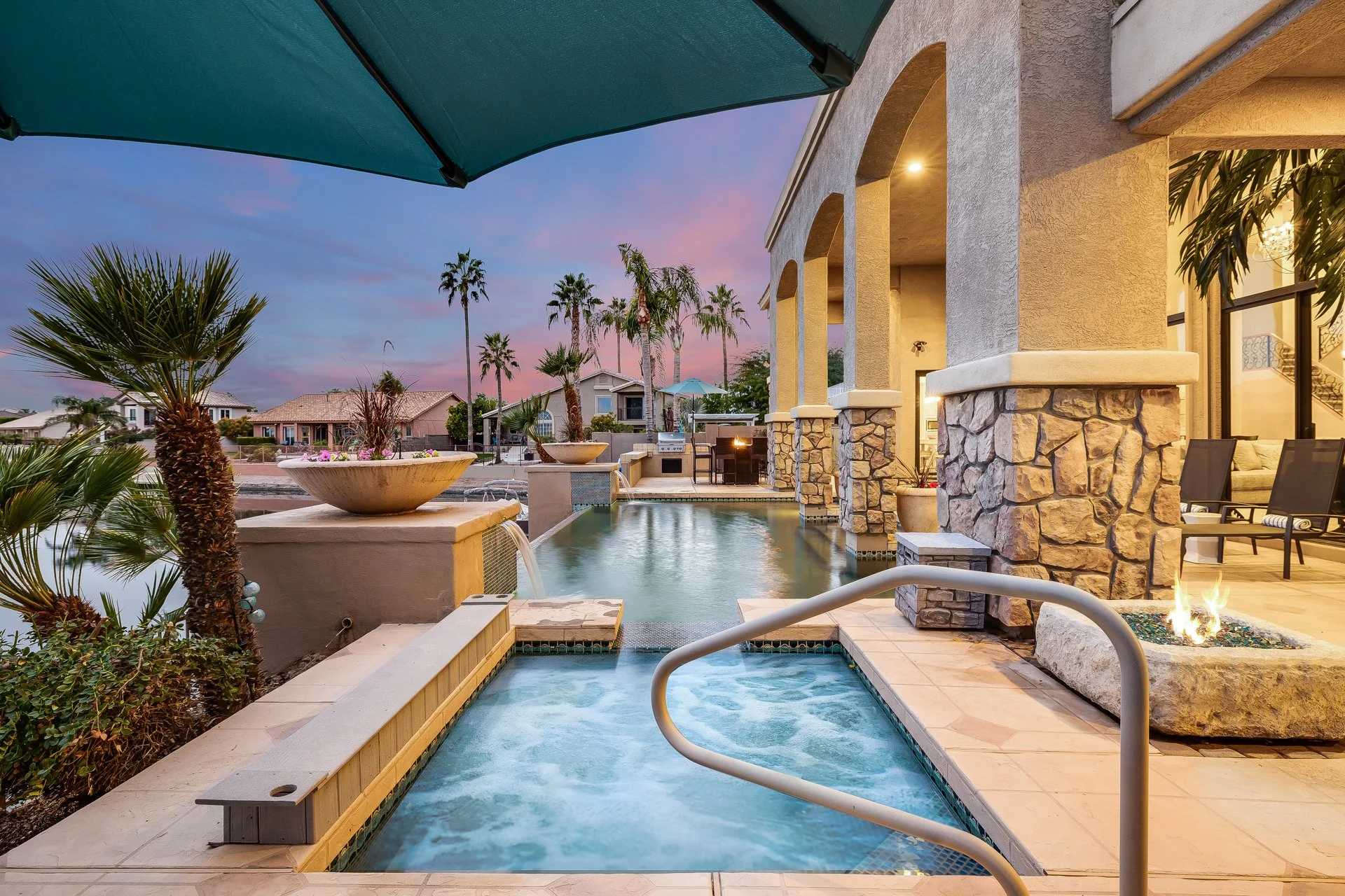 An outdoor swimming pool area at sunset with lounge chairs, palm trees, and a covered patio with stone columns and an outdoor fireplace.