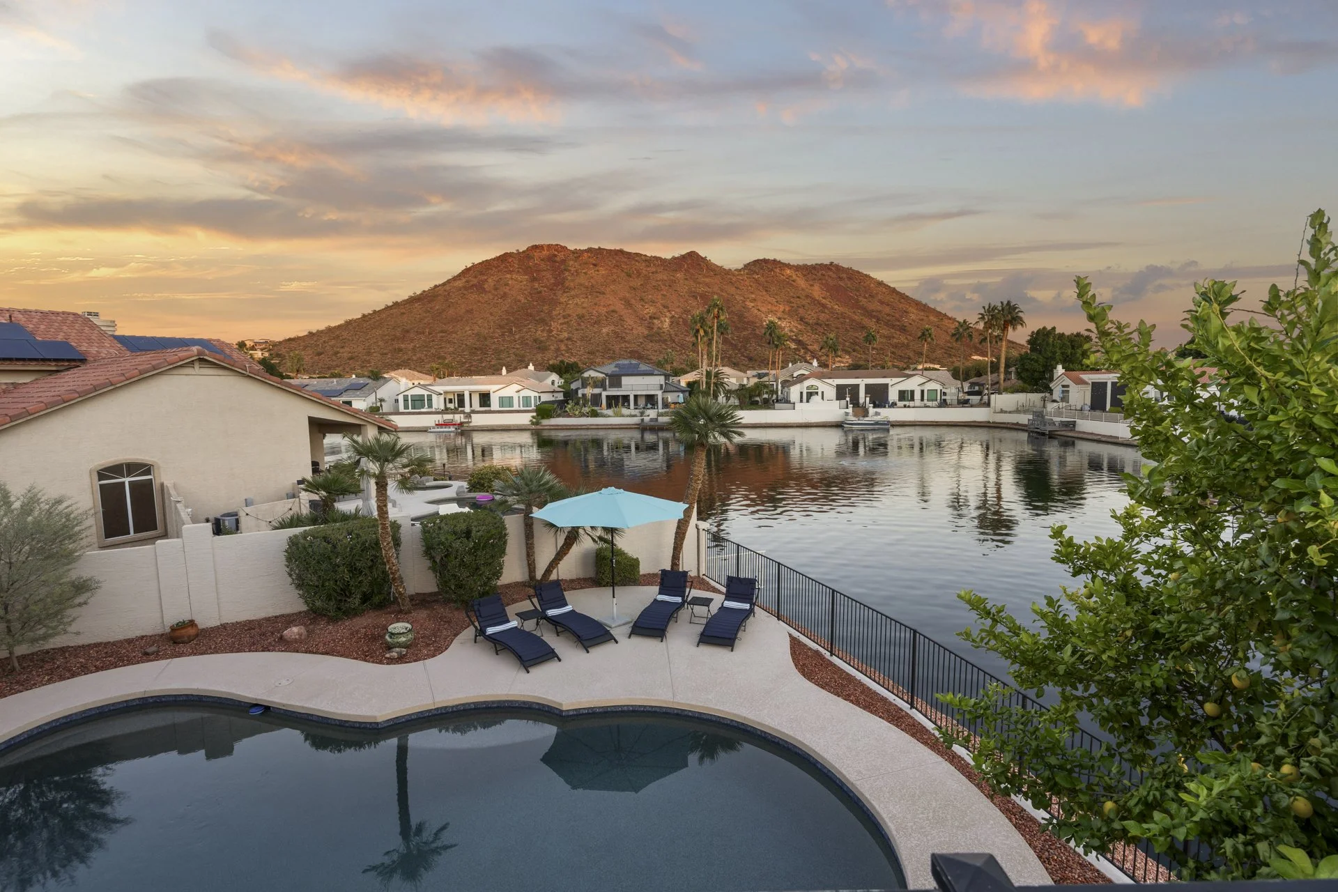 A backyard scene with a swimming pool, a patio with lounge chairs and an umbrella, a waterway with boats, white houses along the water, and a mountain in the background during sunset.
